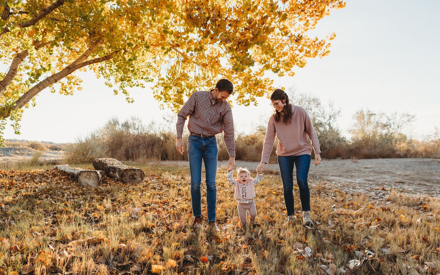 A family walks together holding hands on a path covered in fallen autumn leaves.