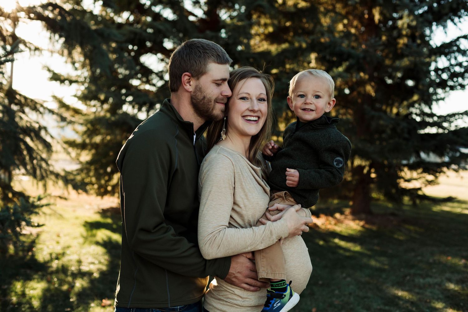 Intimate family portrait showing affectionate parents and toddler against natural forest backdrop.