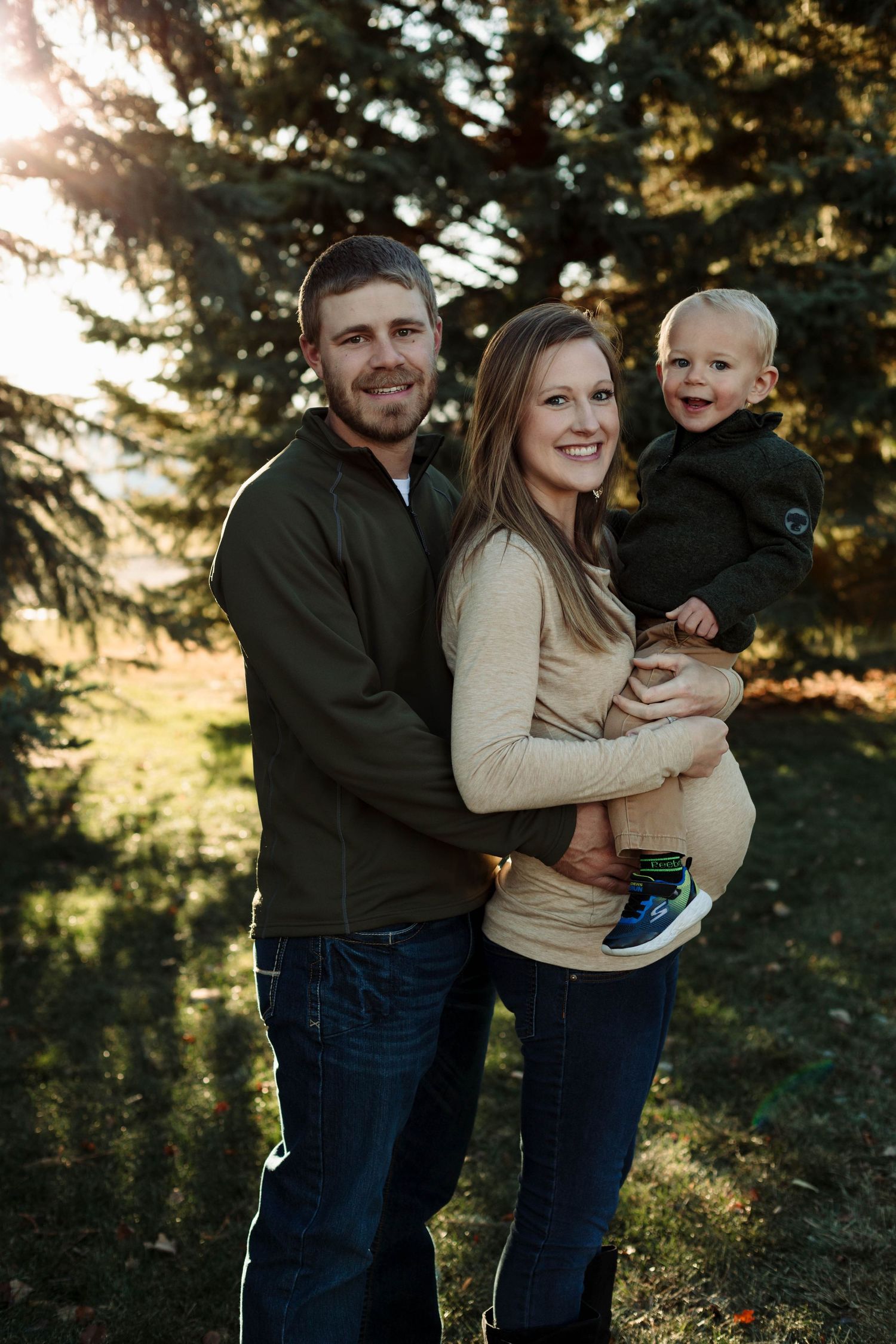 Expectant parents and toddler pose together in front of evergreen trees during golden hour outdoor photo session.