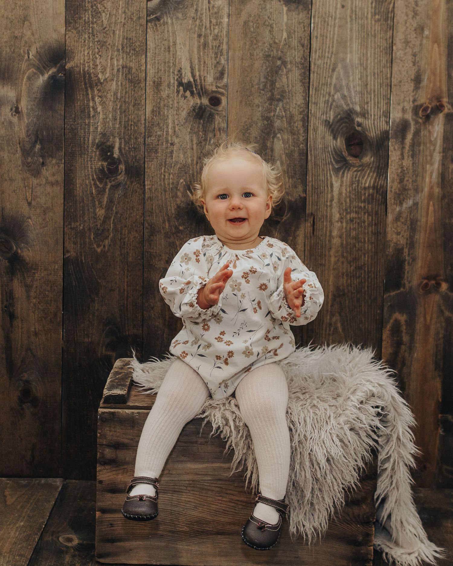 Smiling baby in floral outfit sitting on wooden crate against rustic wall.