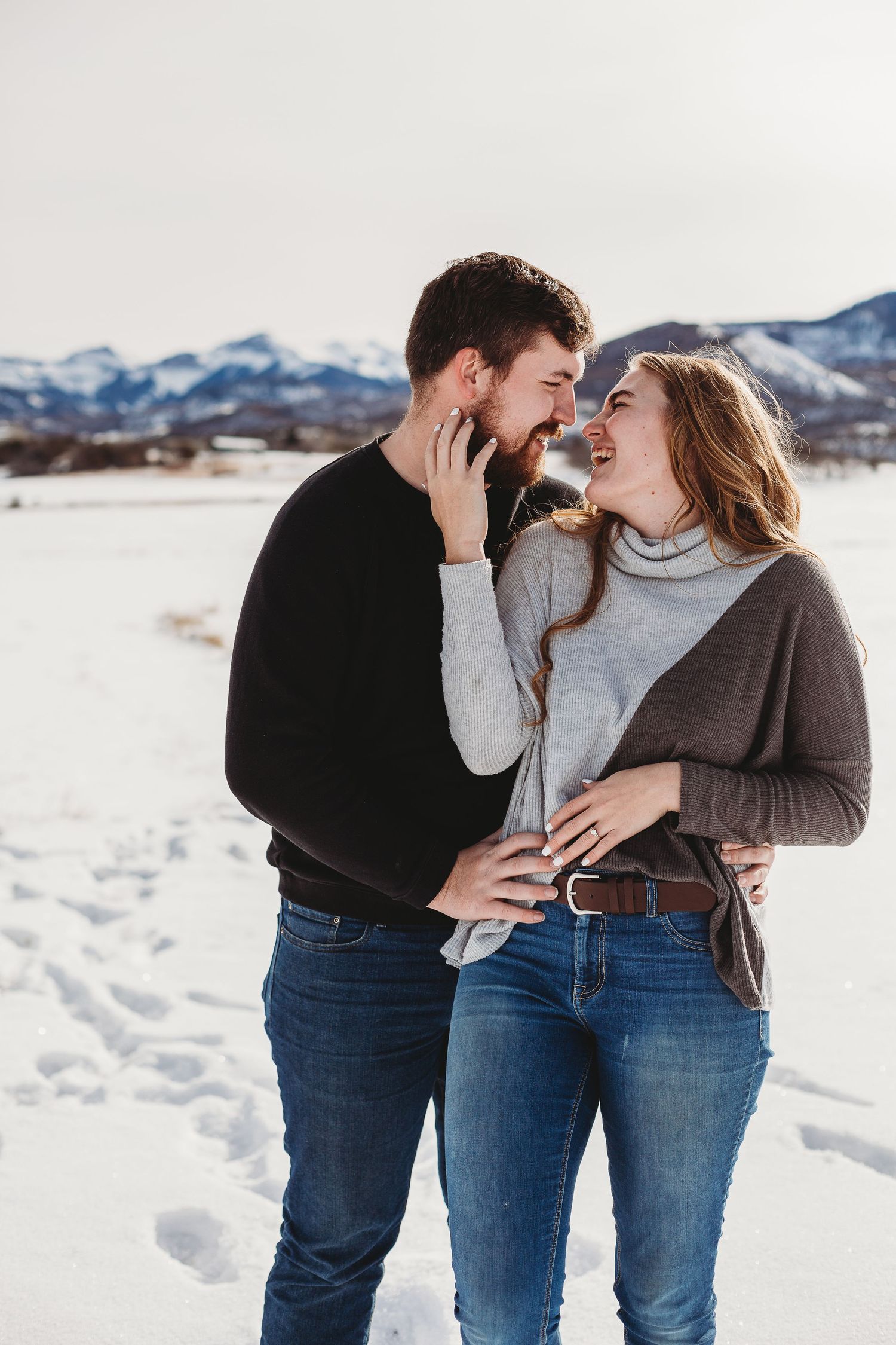 A couple laughs together while embracing in a snowy mountain setting.