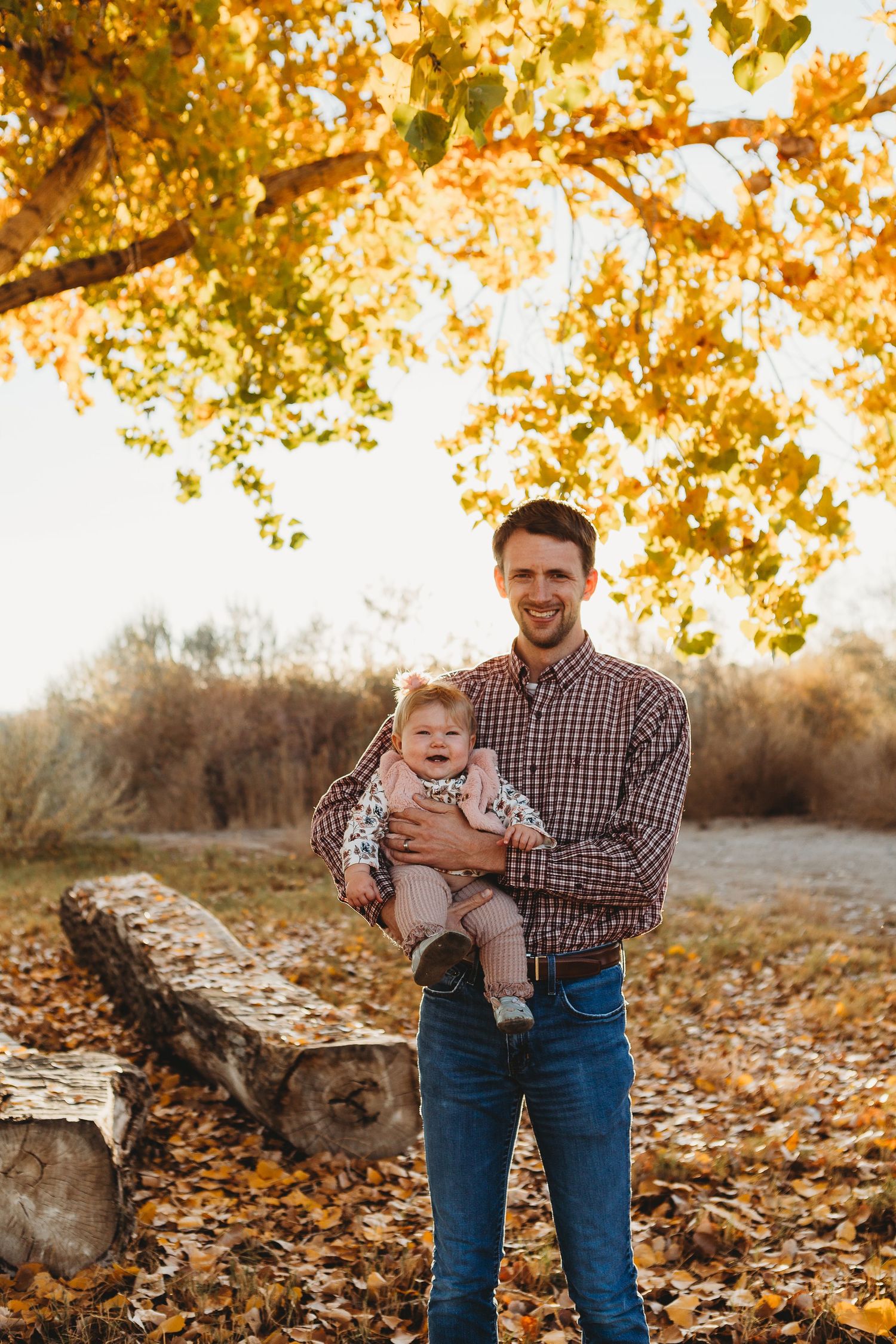 A parent holds a smiling baby under golden autumn trees during an outdoor portrait session.