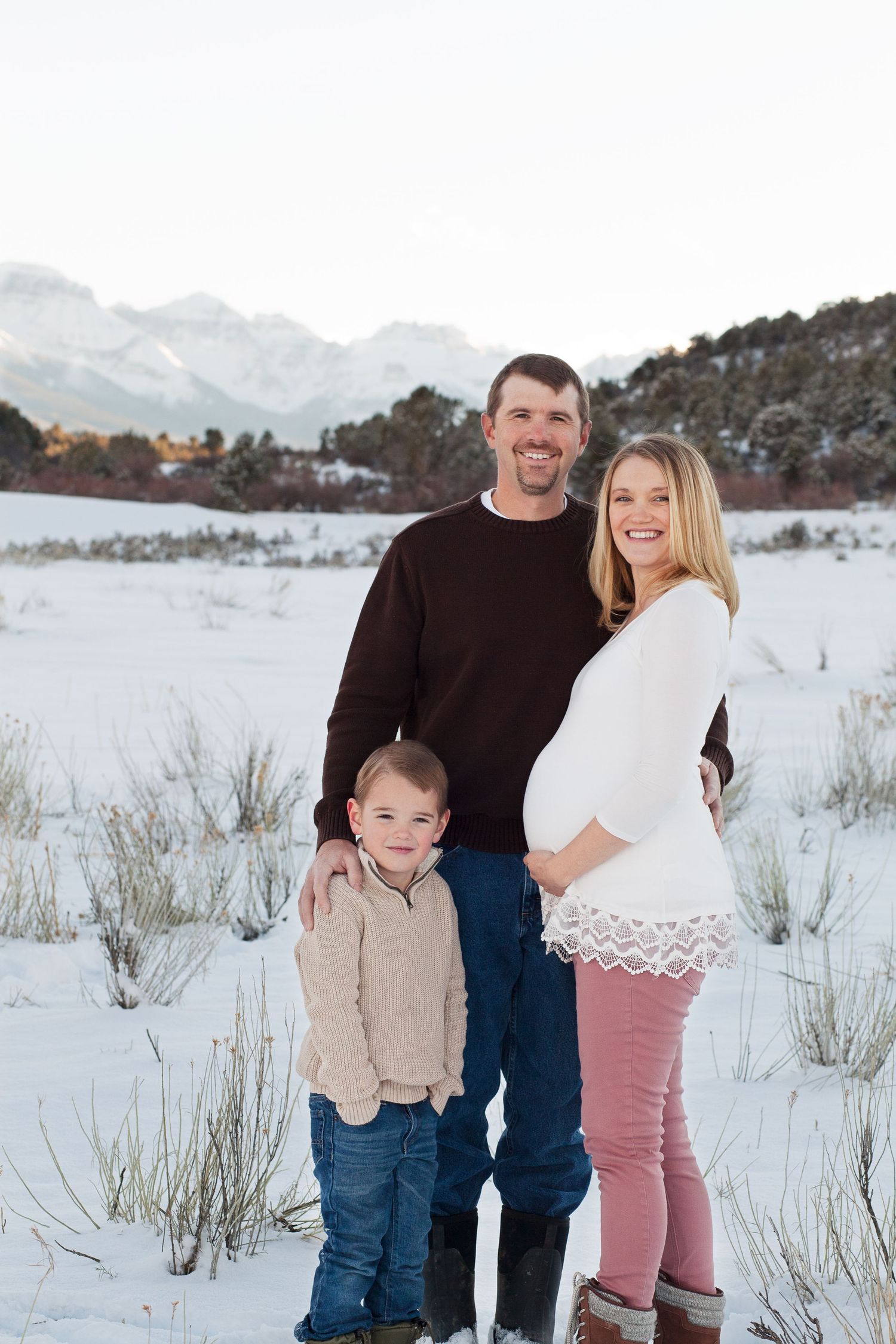 Family of three poses together in snowy mountain landscape during winter maternity shoot.