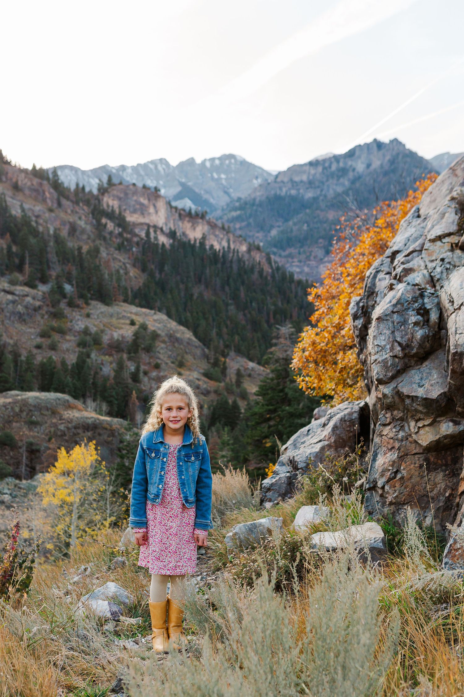 Child in denim jacket and dress standing among fall foliage with mountain backdrop.