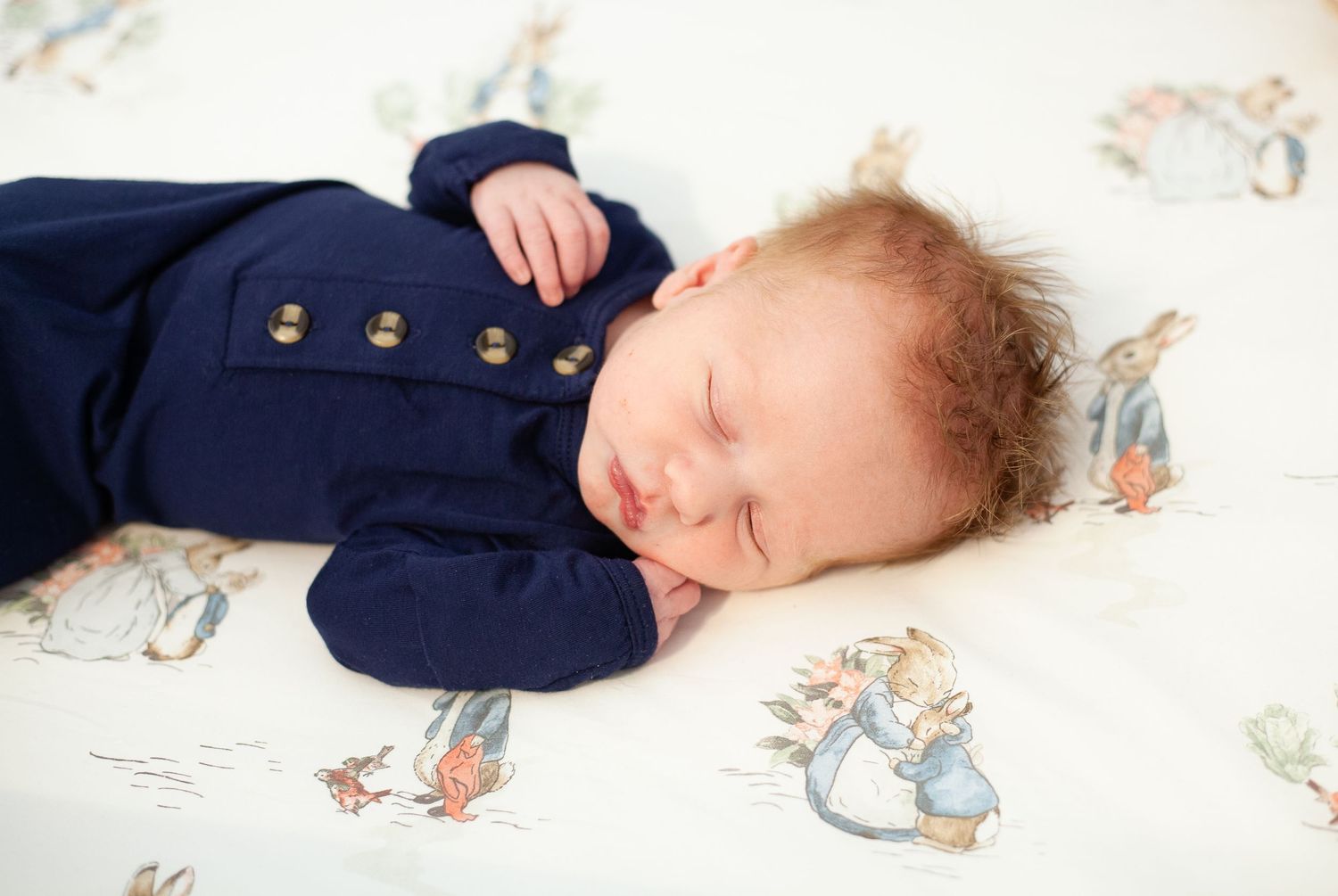 A sleeping baby in a navy blue outfit rests on Peter Rabbit patterned bedding.