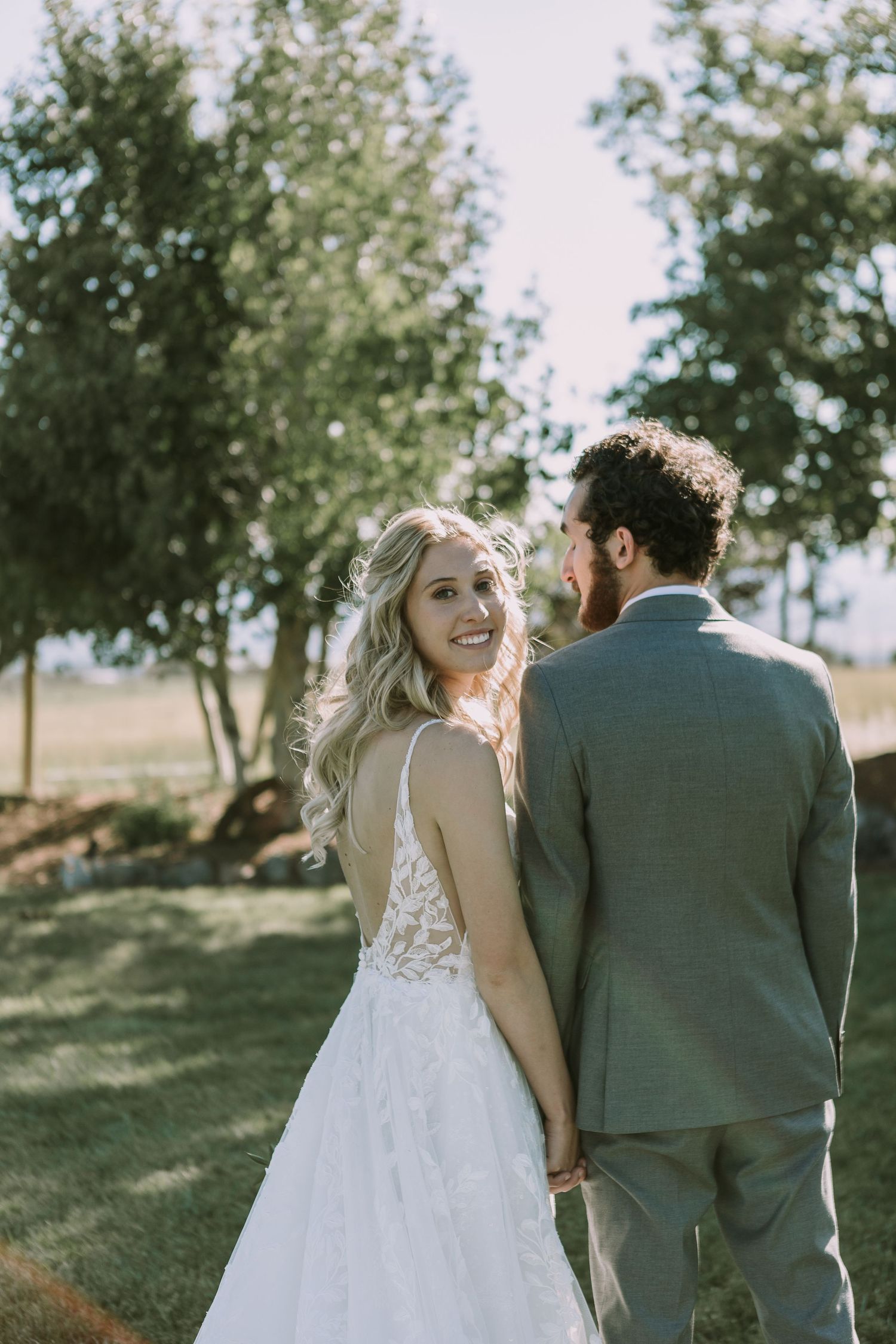 A bride in a lace wedding dress looks back at her groom in a gray suit under trees in natural light.