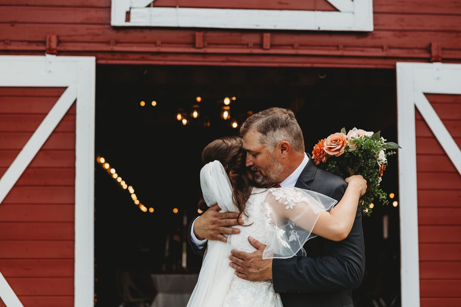 Sweet embrace in front of red barn doors with twinkling lights.