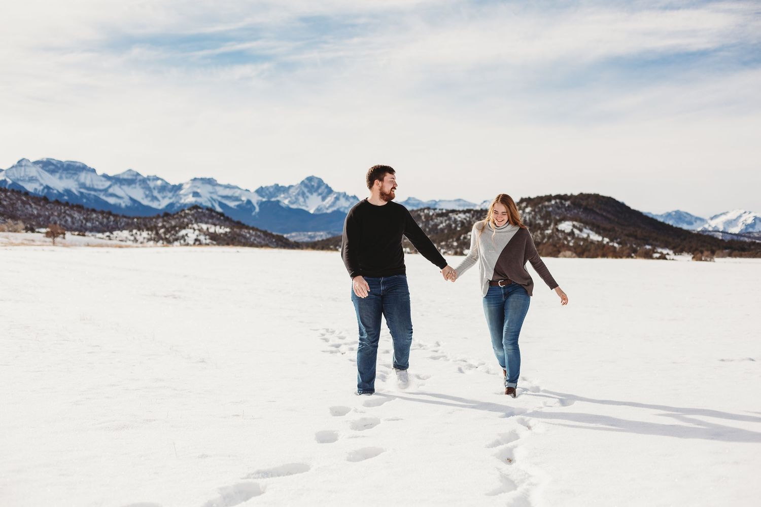 A joyful couple playing and jumping in the snow with snow-capped mountains behind them.
