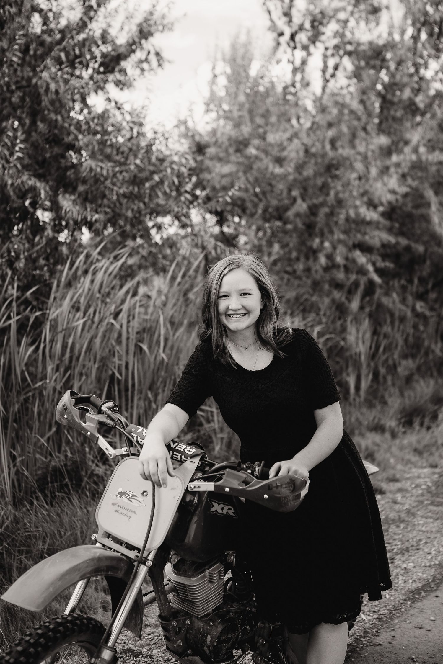 Person in black dress poses with dirt bike in front of natural foliage backdrop.