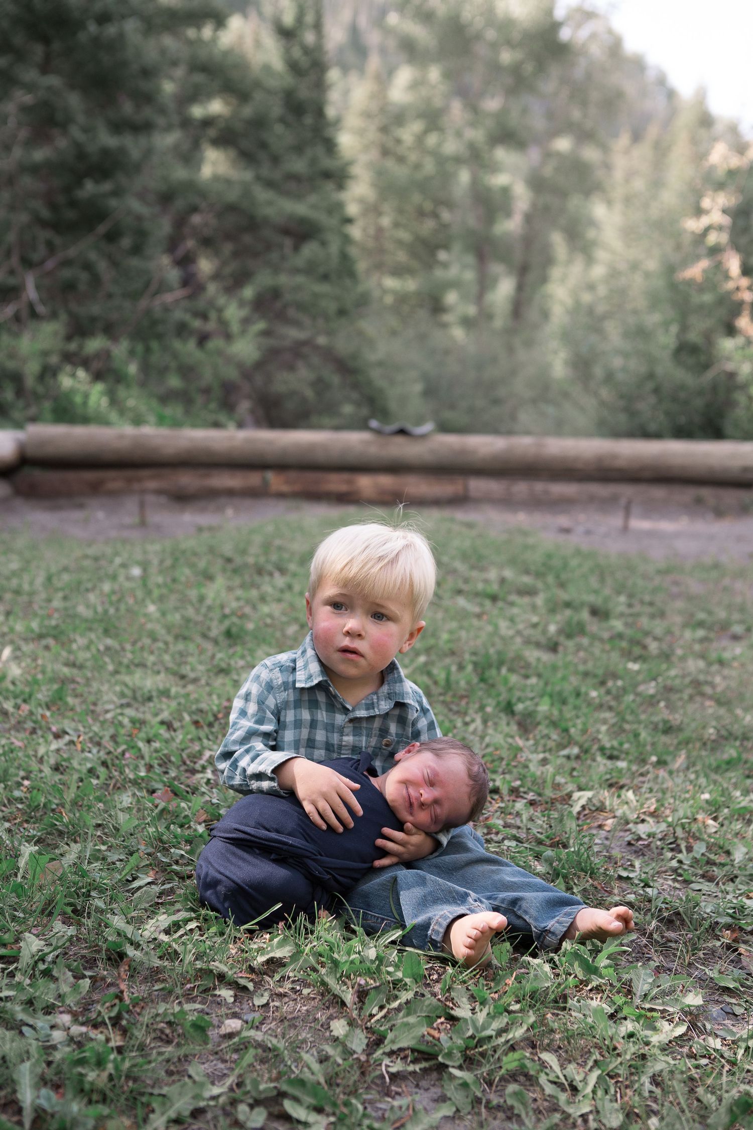 Two siblings share a tender moment while sitting on grass in a mountain setting.