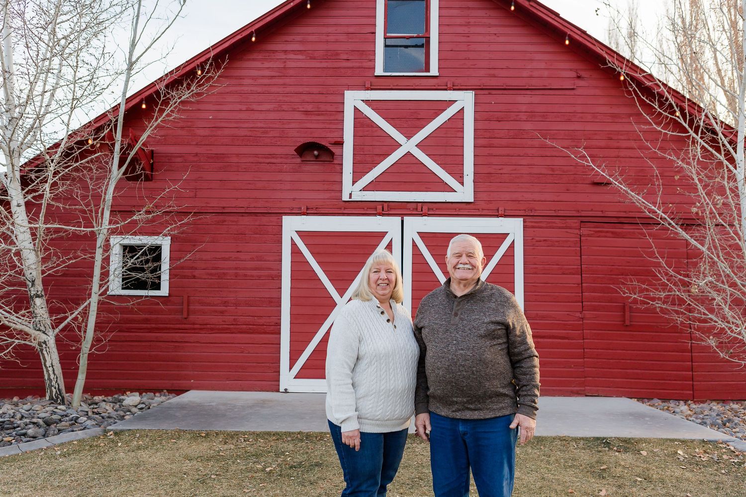 A couple stands together in front of a classic red barn with white X-patterned doors.