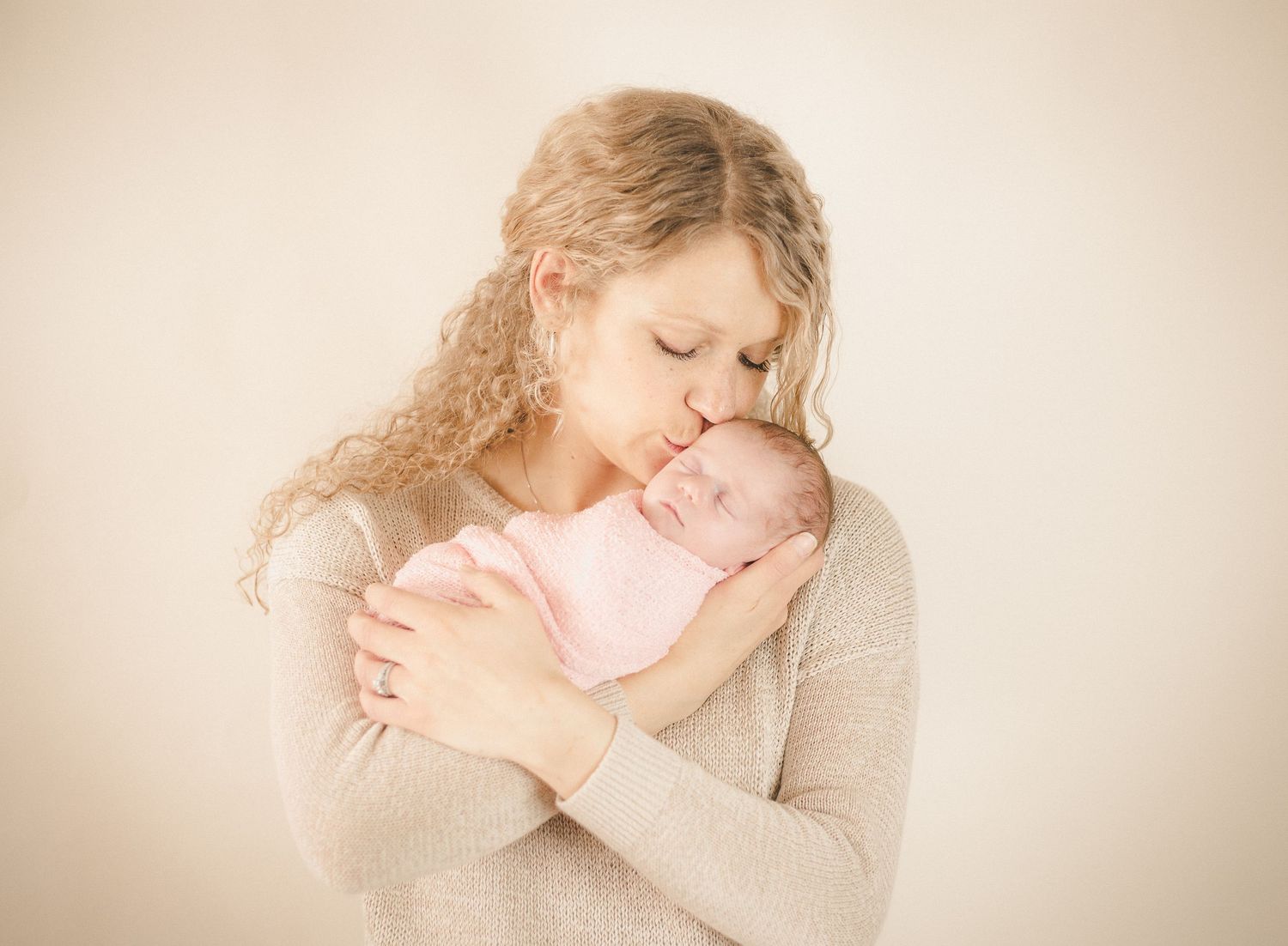 Gentle maternal moment captured in soft lighting against beige background.