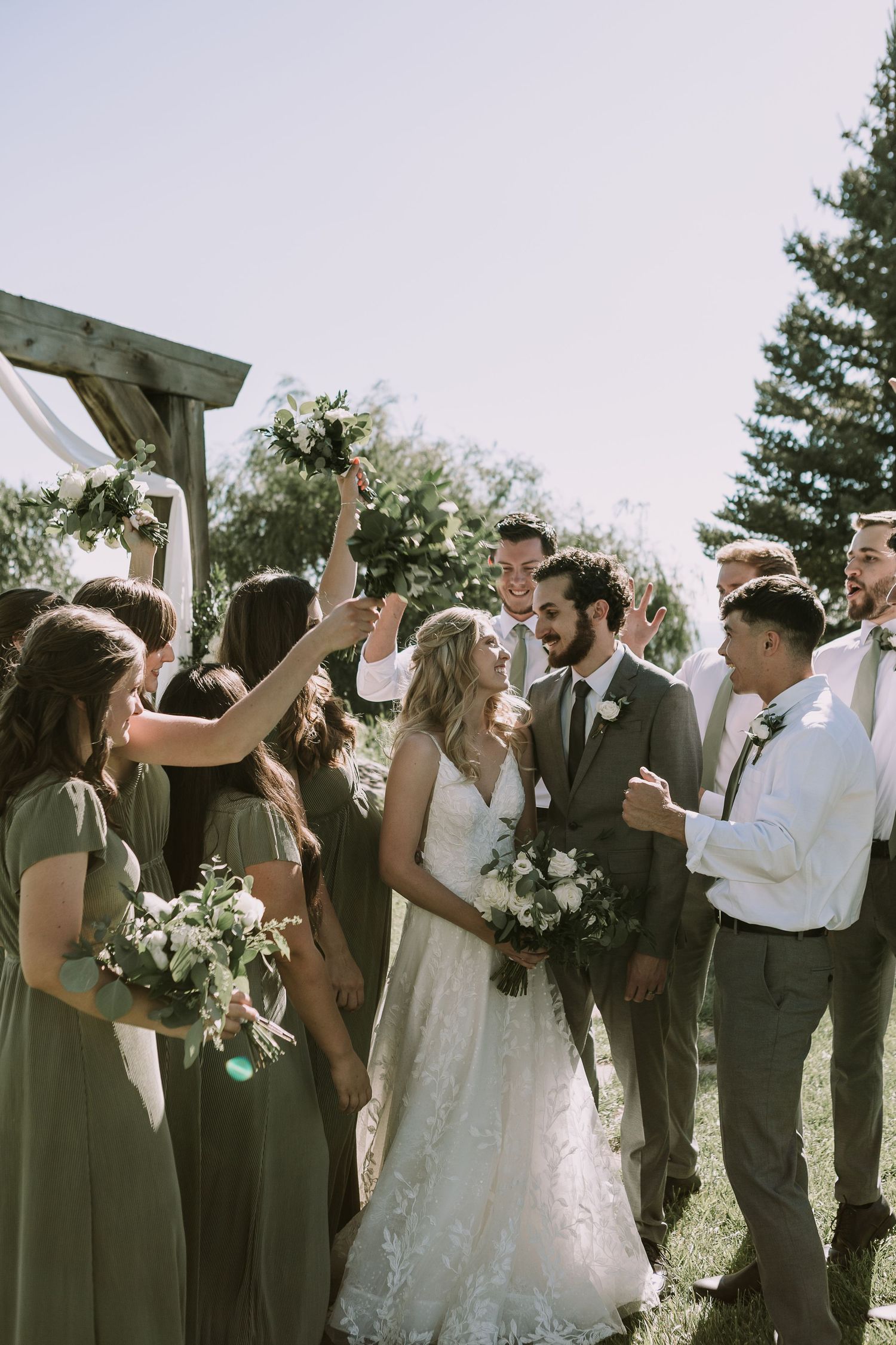 Wedding party cheers and celebrates around newlyweds under wooden arch outdoors.