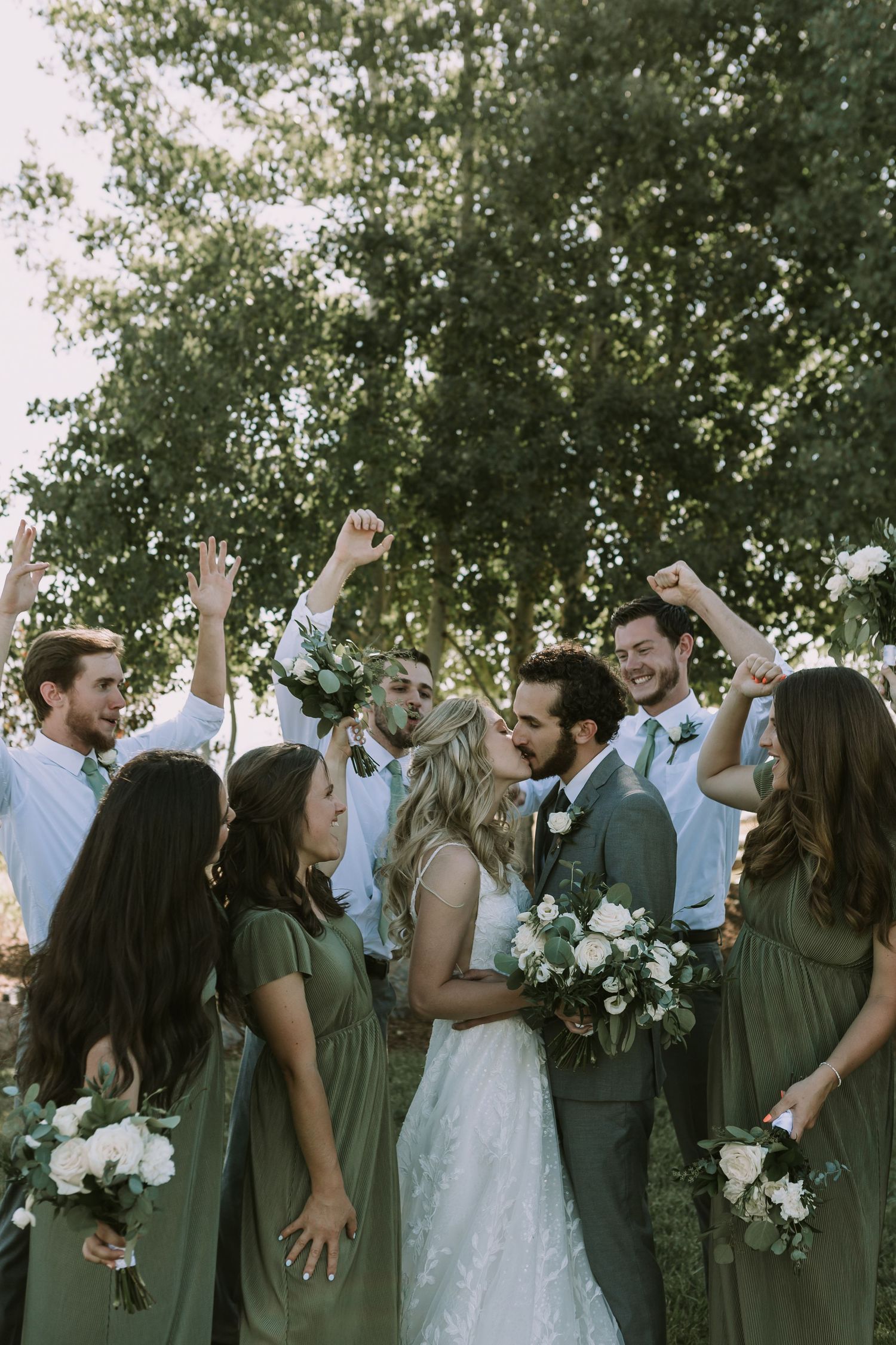 Joyful wedding party celebrates with raised arms among trees.