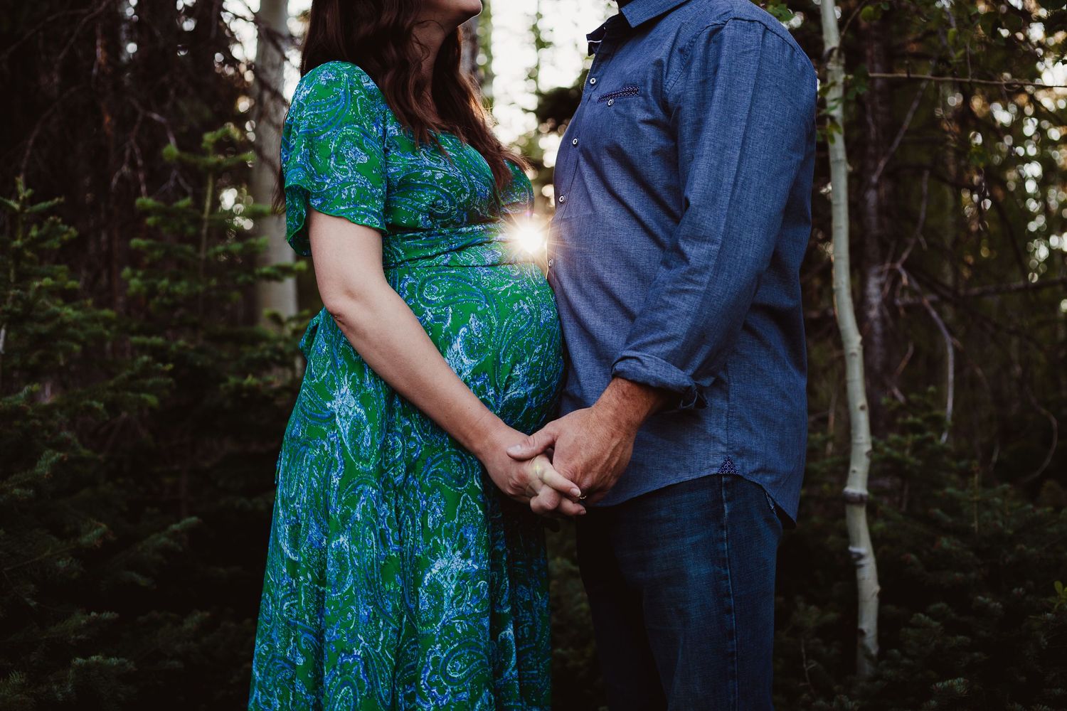 A backlit maternity photo of a couple holding hands among birch trees at sunset.