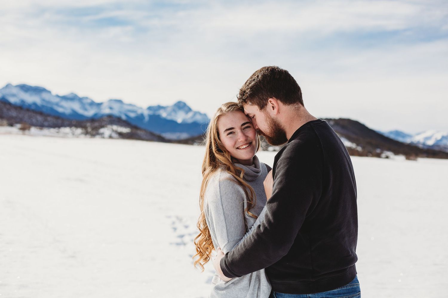 A loving couple embraces in a winter setting with snow-capped mountains in the background.