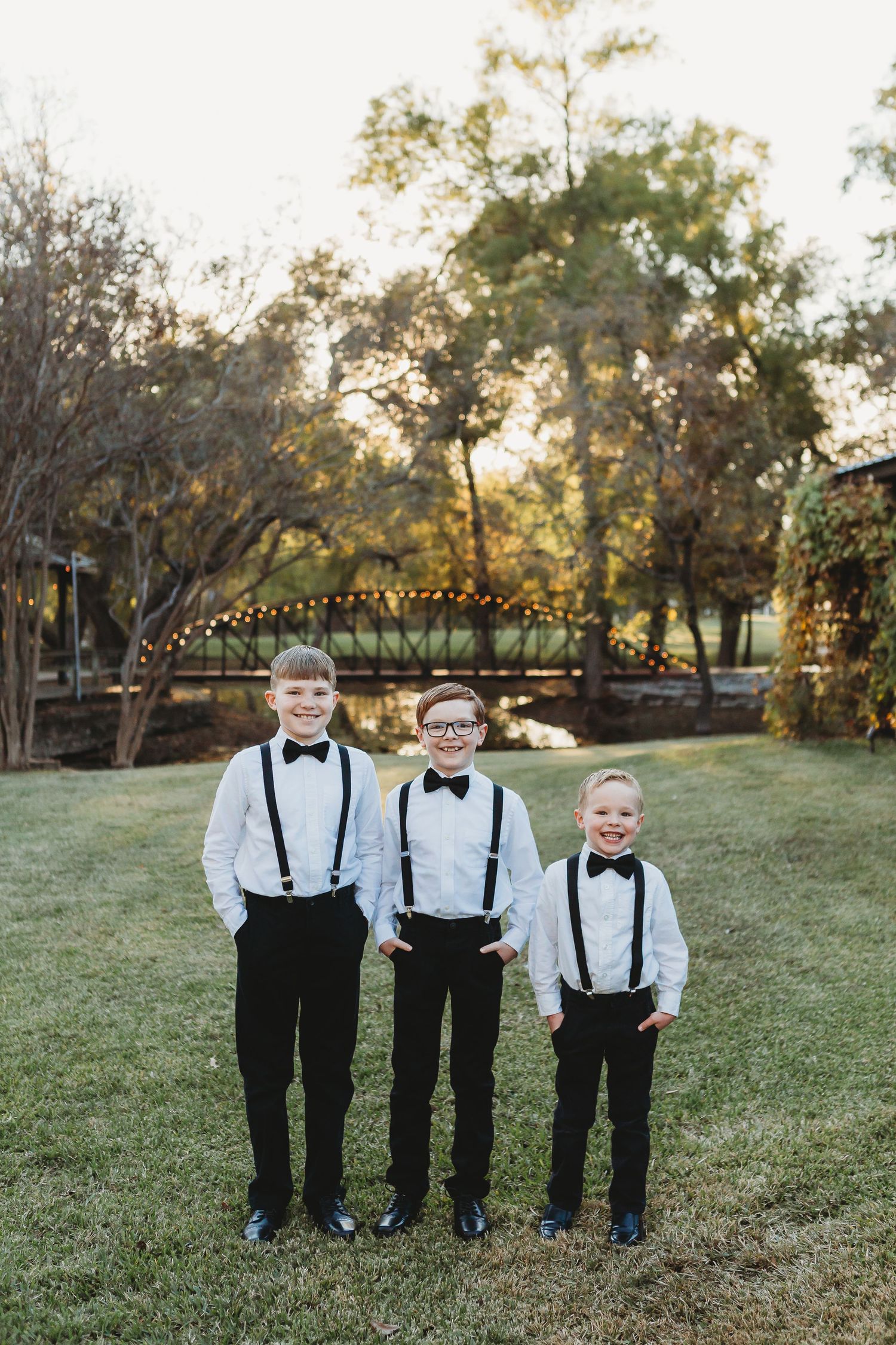 Young groomsmen in formal black suits with suspenders and bowties stand together by wooden bridge.