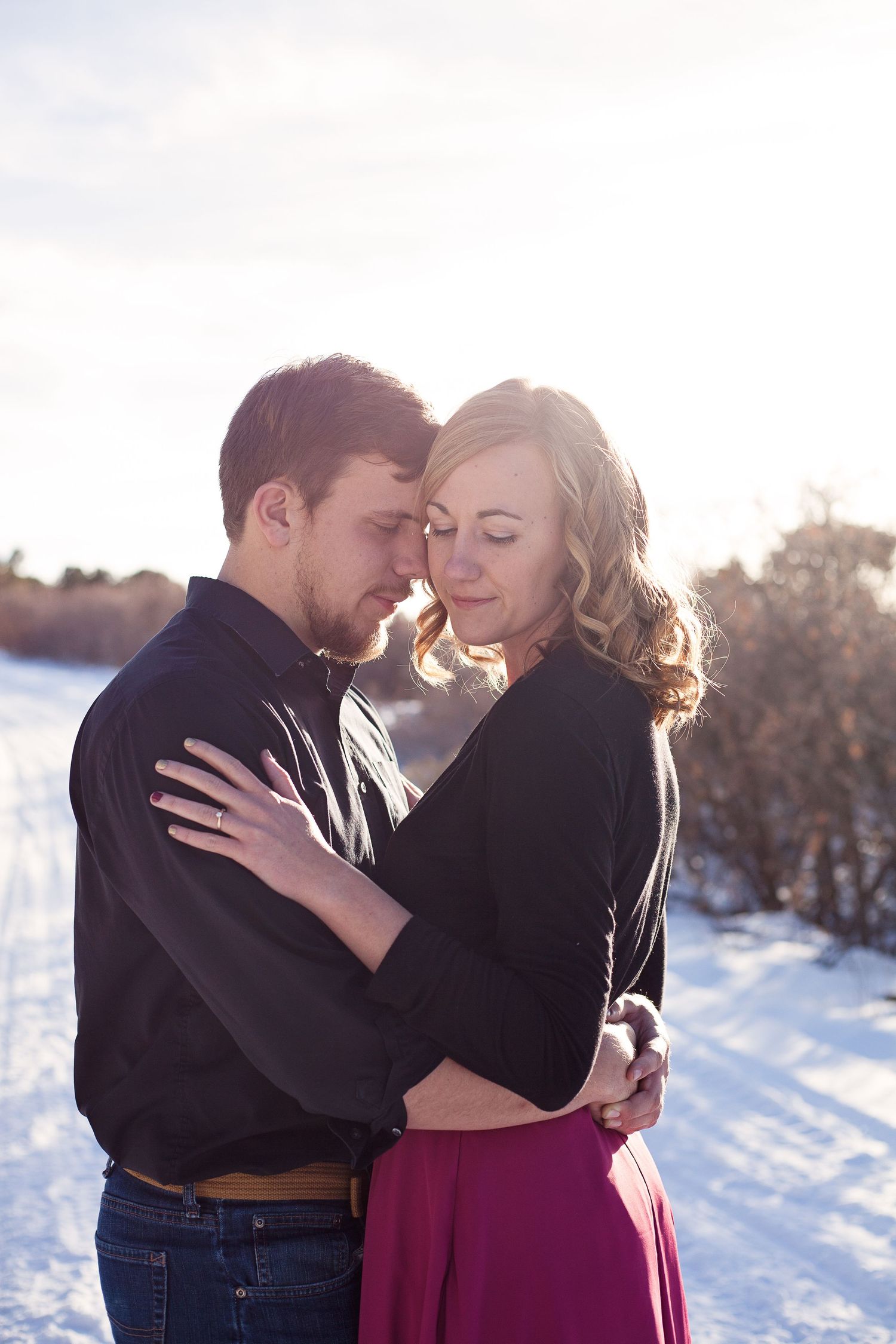 A romantic couple embraces in winter sunlight against a snowy backdrop.