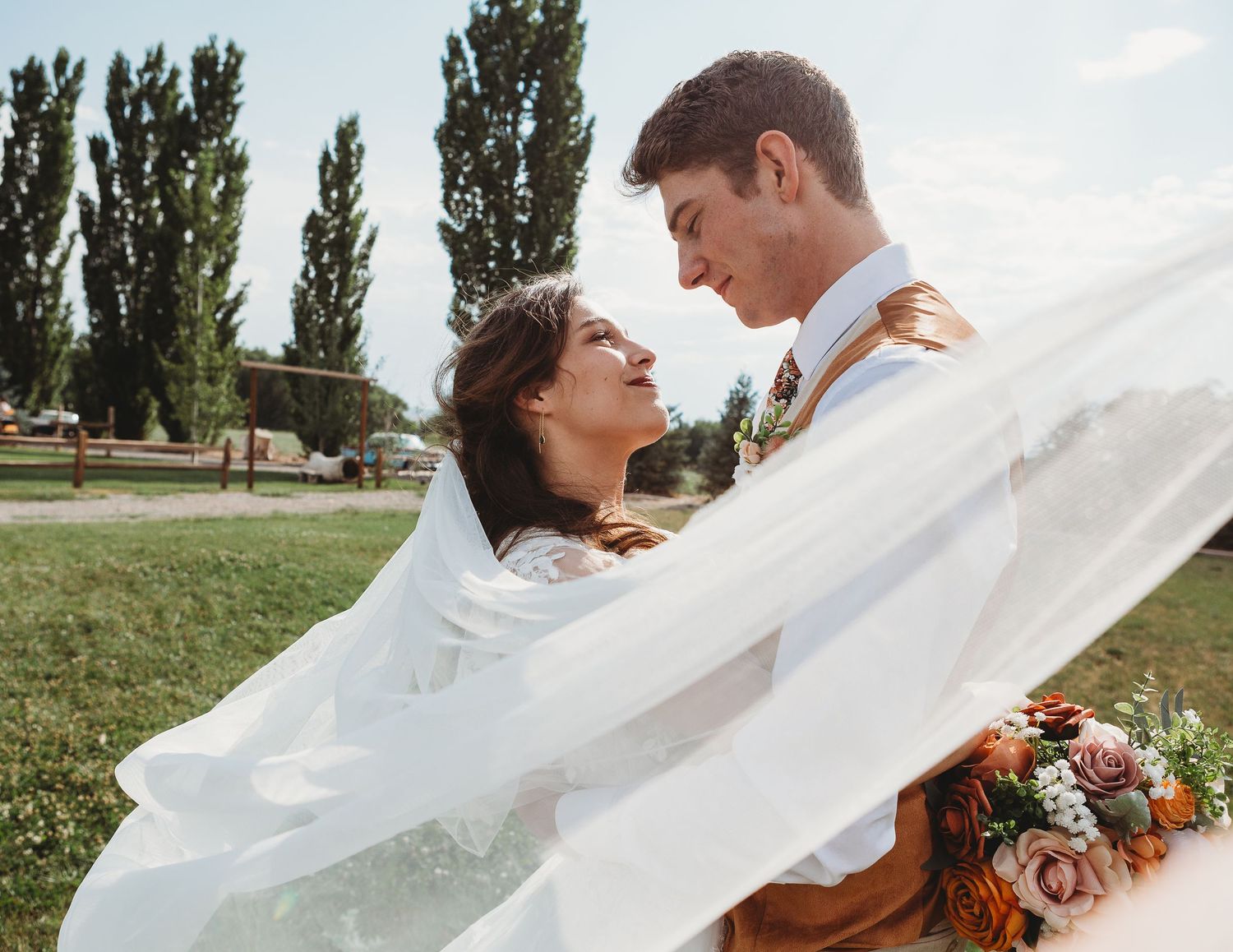 Wedding couple embraces outdoors as veil flows dramatically in the wind.