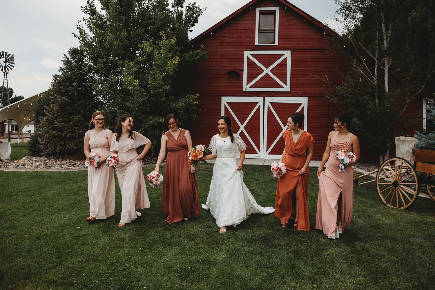 Bridal party in coordinating rust and blush dresses poses on lawn with red barn backdrop.