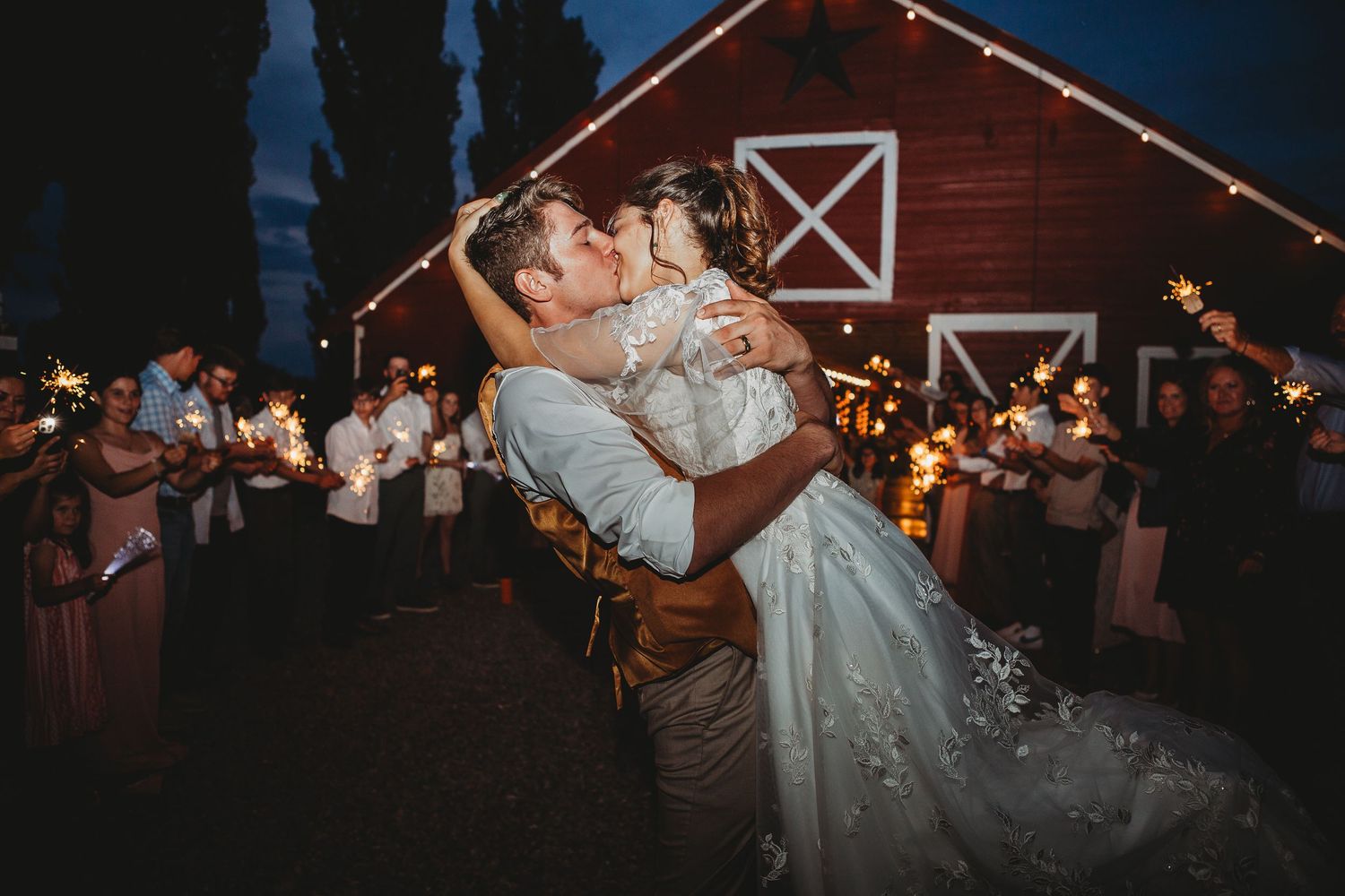 Bride and groom share romantic moment outside illuminated red barn at night.