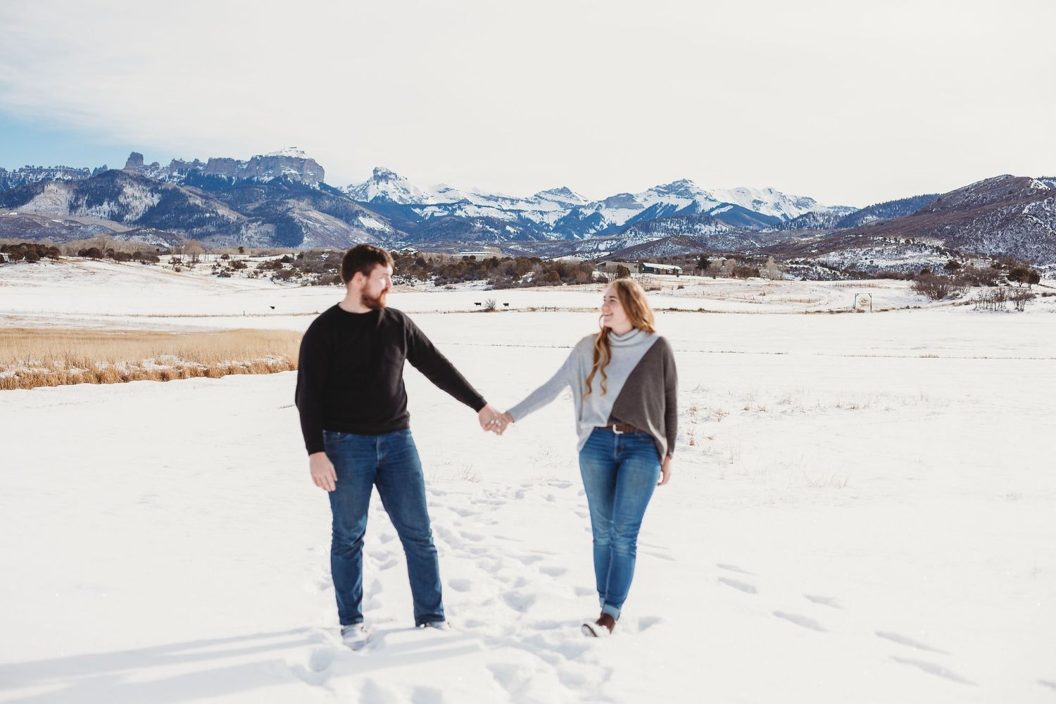 A couple holds hands while walking through a snow-covered field with mountains in the distance.