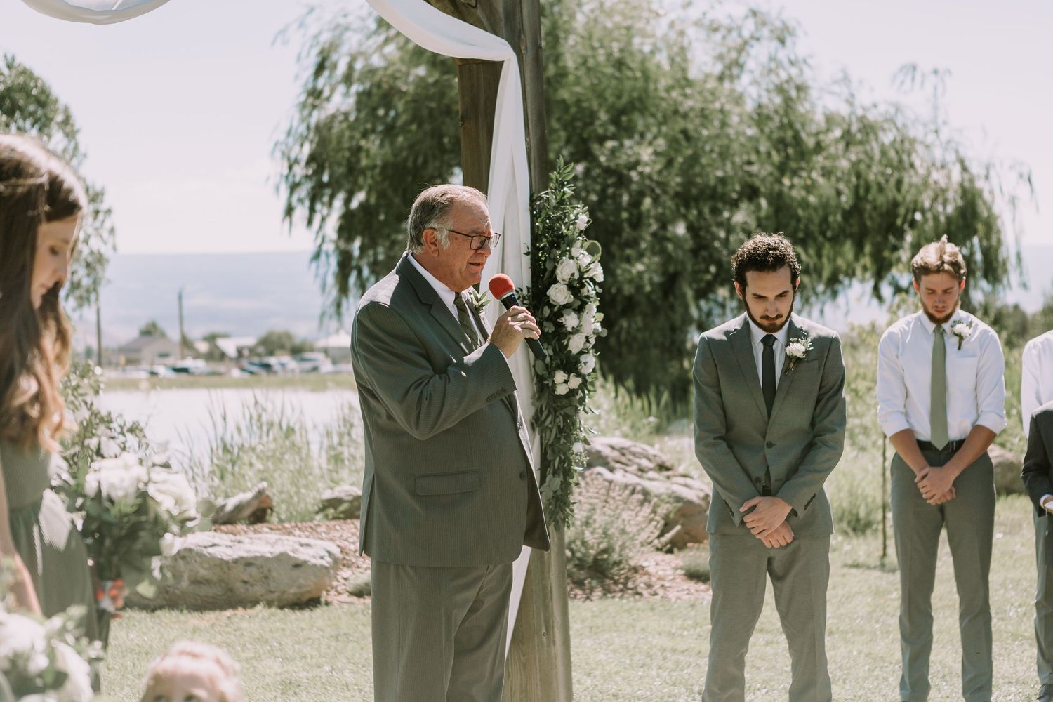Outdoor wedding ceremony with officiant speaking at rustic wooden arch draped in white fabric and florals.