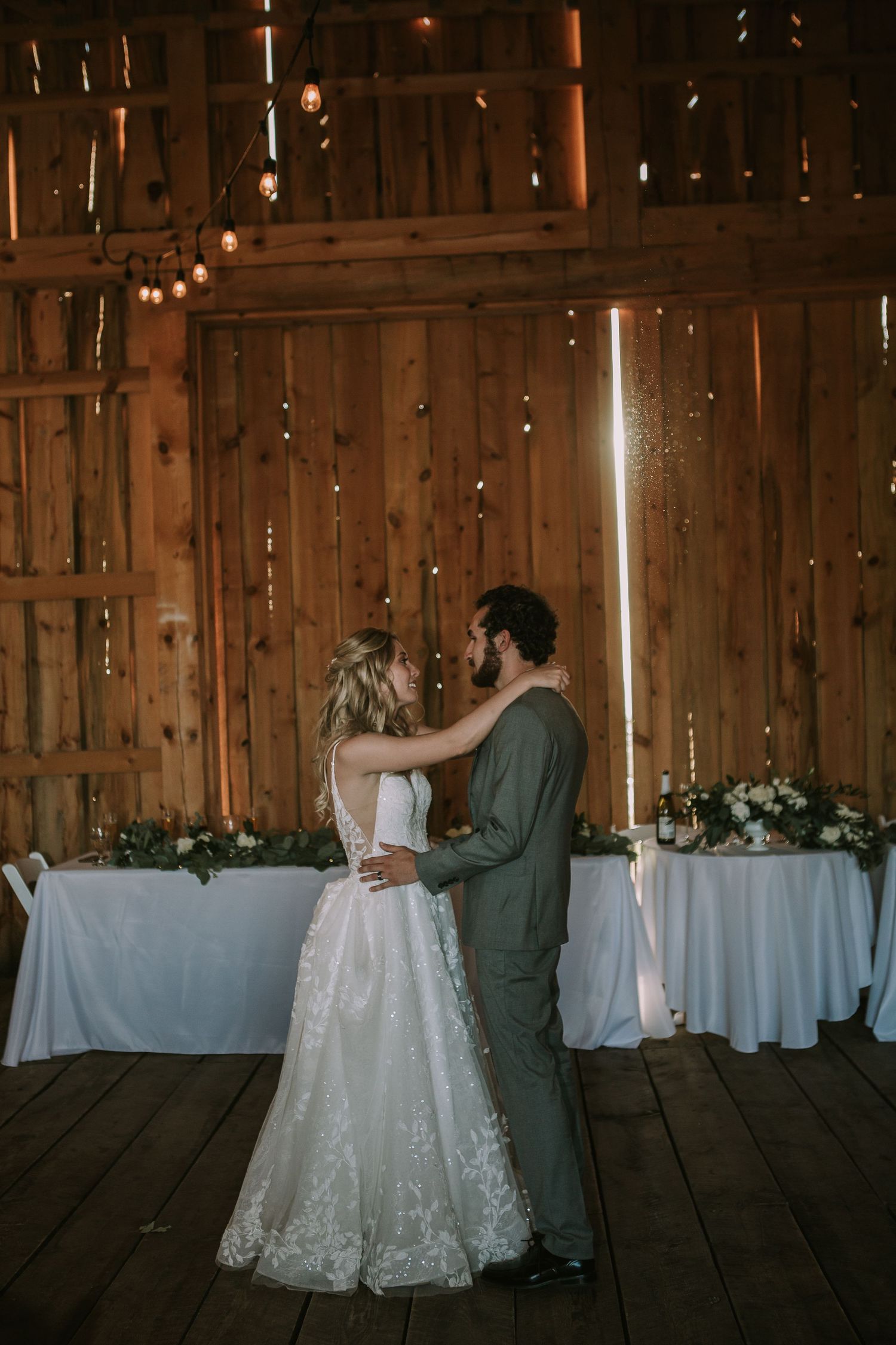 Romantic first dance moment in a dimly lit rustic barn with string lights and white table settings.