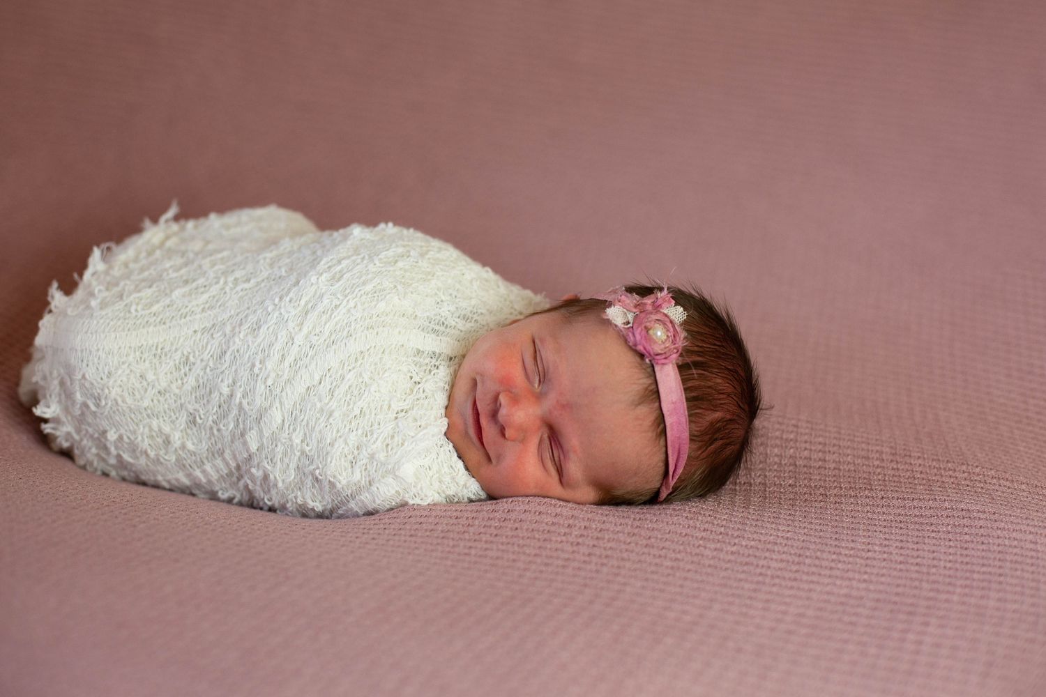 Sleeping newborn wrapped in white textured blanket with pink headband.