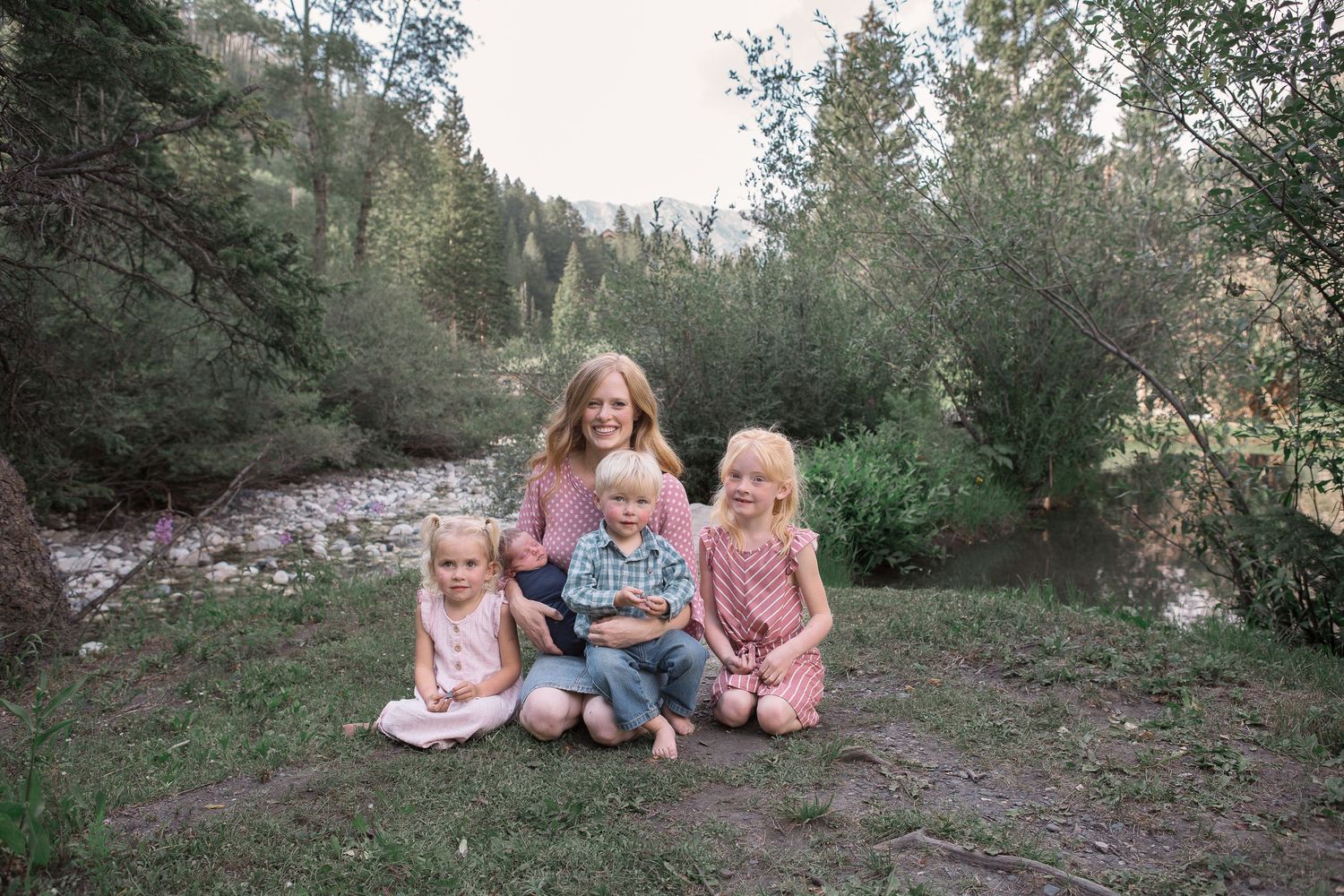 Family group sitting together on grassy ground near a mountain stream surrounded by greenery.