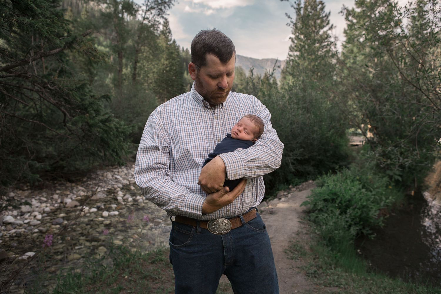 A person in plaid shirt gently cradles a sleeping newborn on a wooded mountain path.