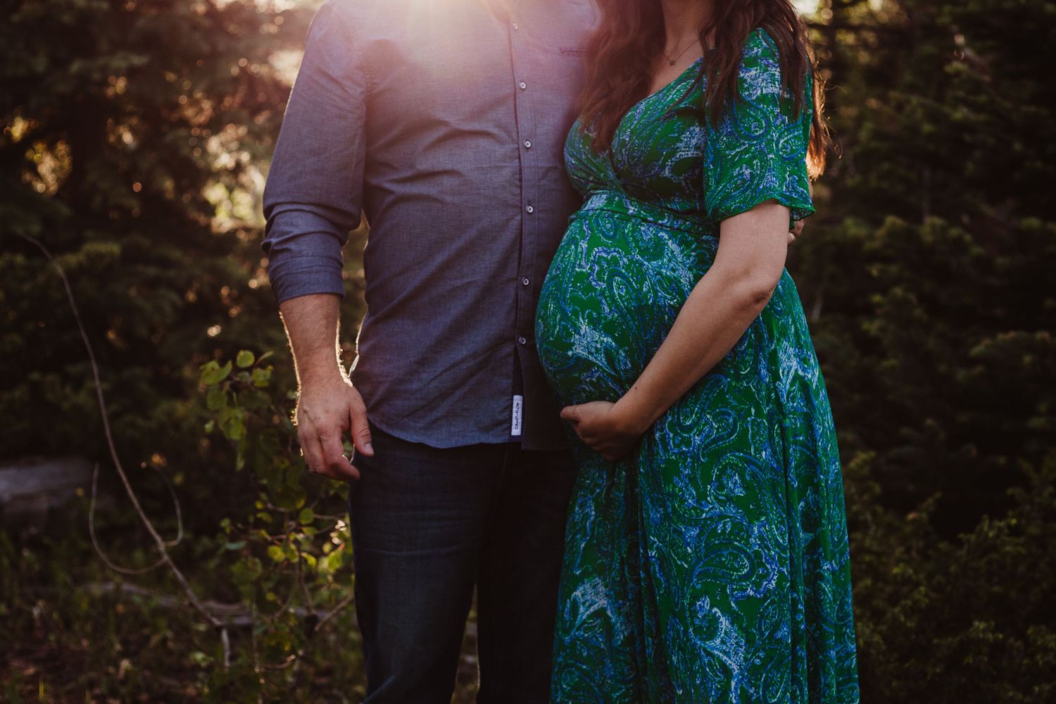 Silhouetted couple in maternity pose against sunset through trees.