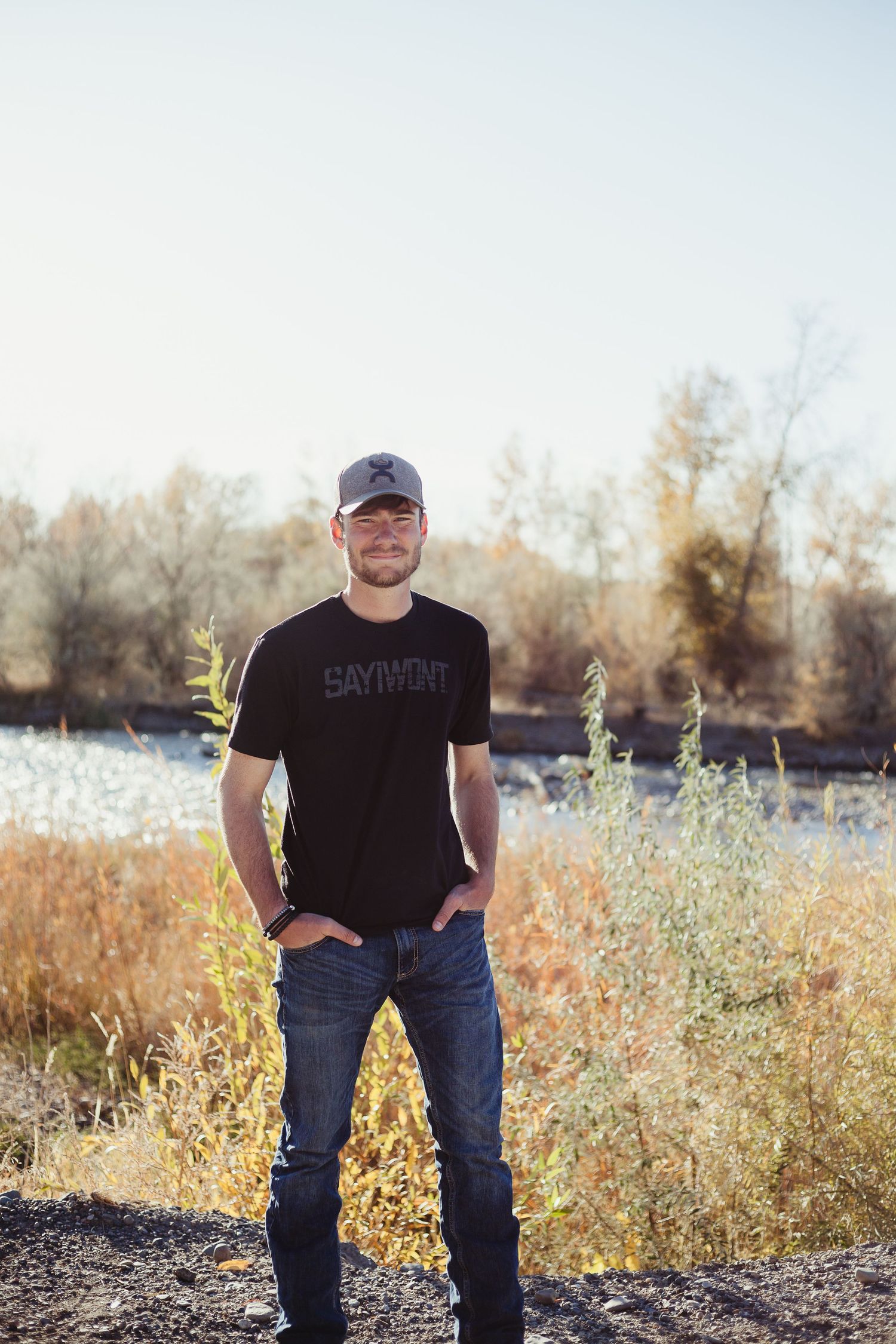 Person in black t-shirt and jeans standing near river with autumn trees in background.
