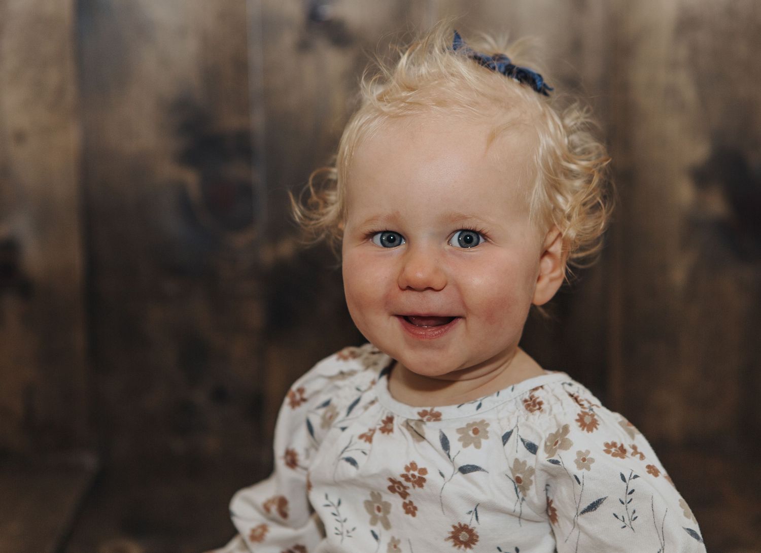 Toddler with curly blonde hair smiling against rustic wooden background.