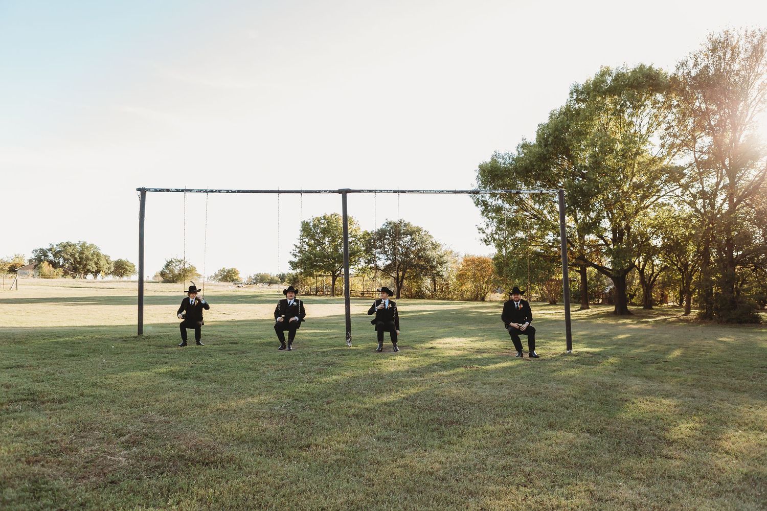 Four people sitting on swings at sunset in a open field with trees in the background.