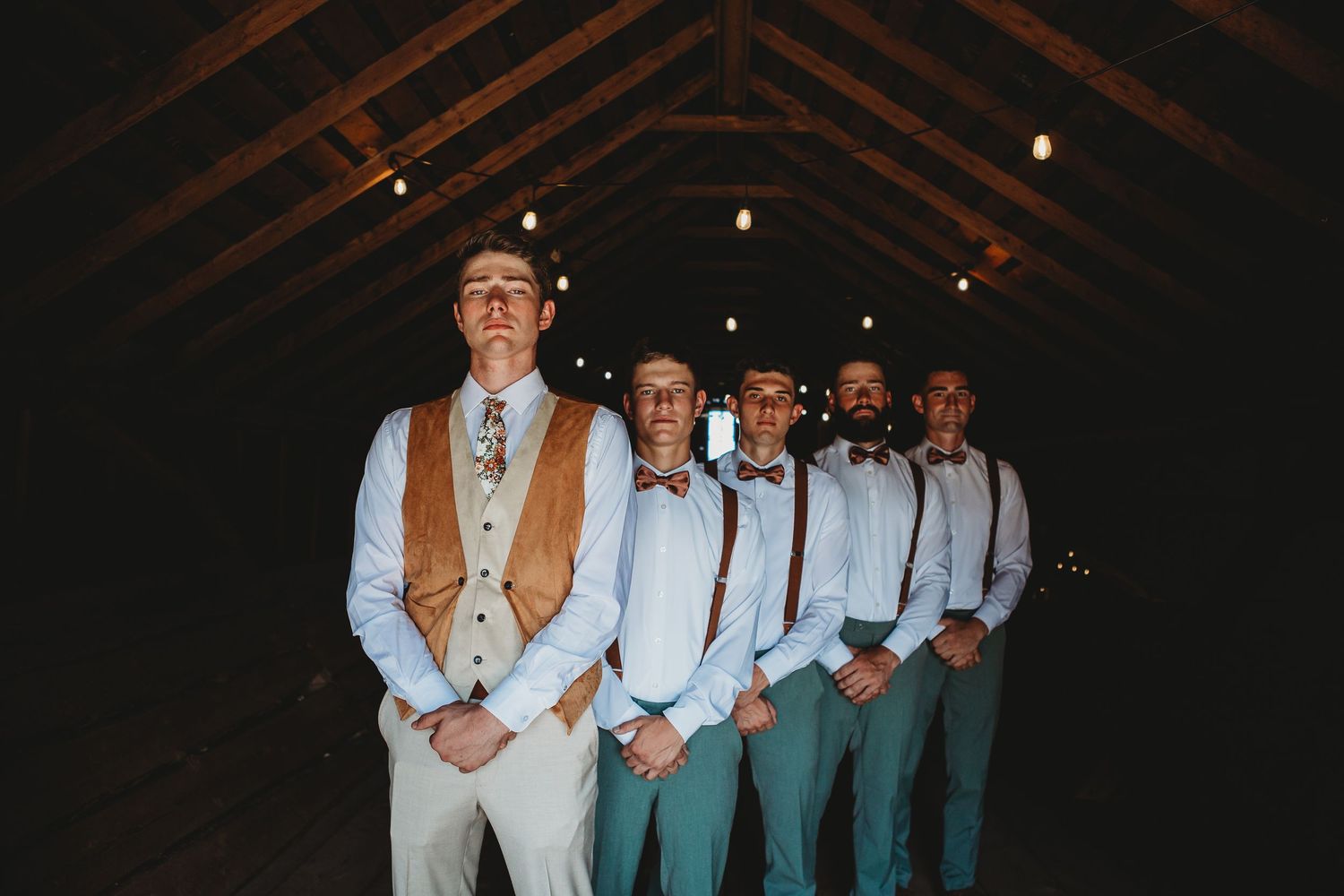 Groomsmen lined up in a rustic barn wearing tan vests and green pants.