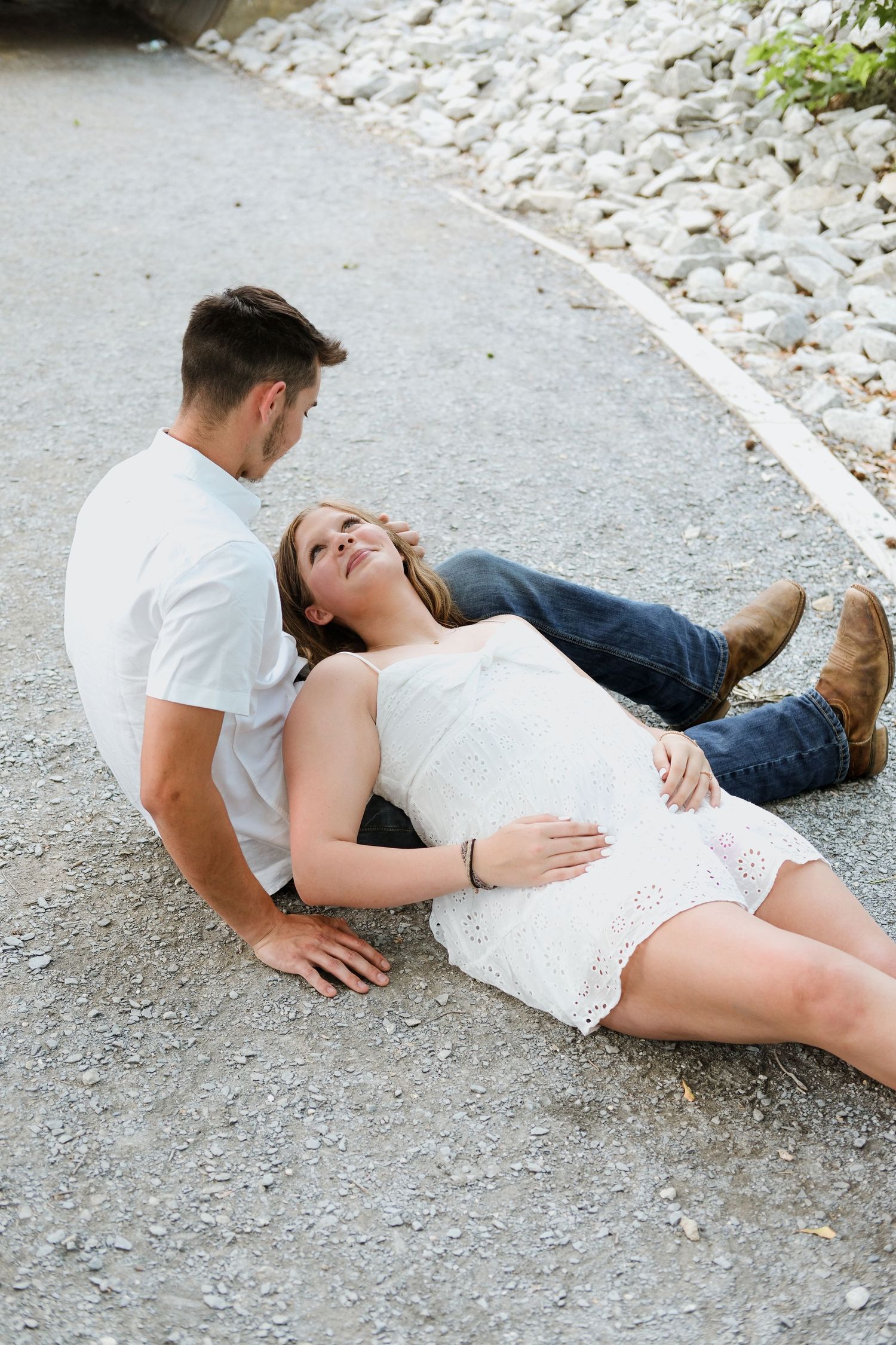 Pregnant couple relaxing on gravel path, woman in white lace dress resting head on mans lap, sharing a tender moment.
