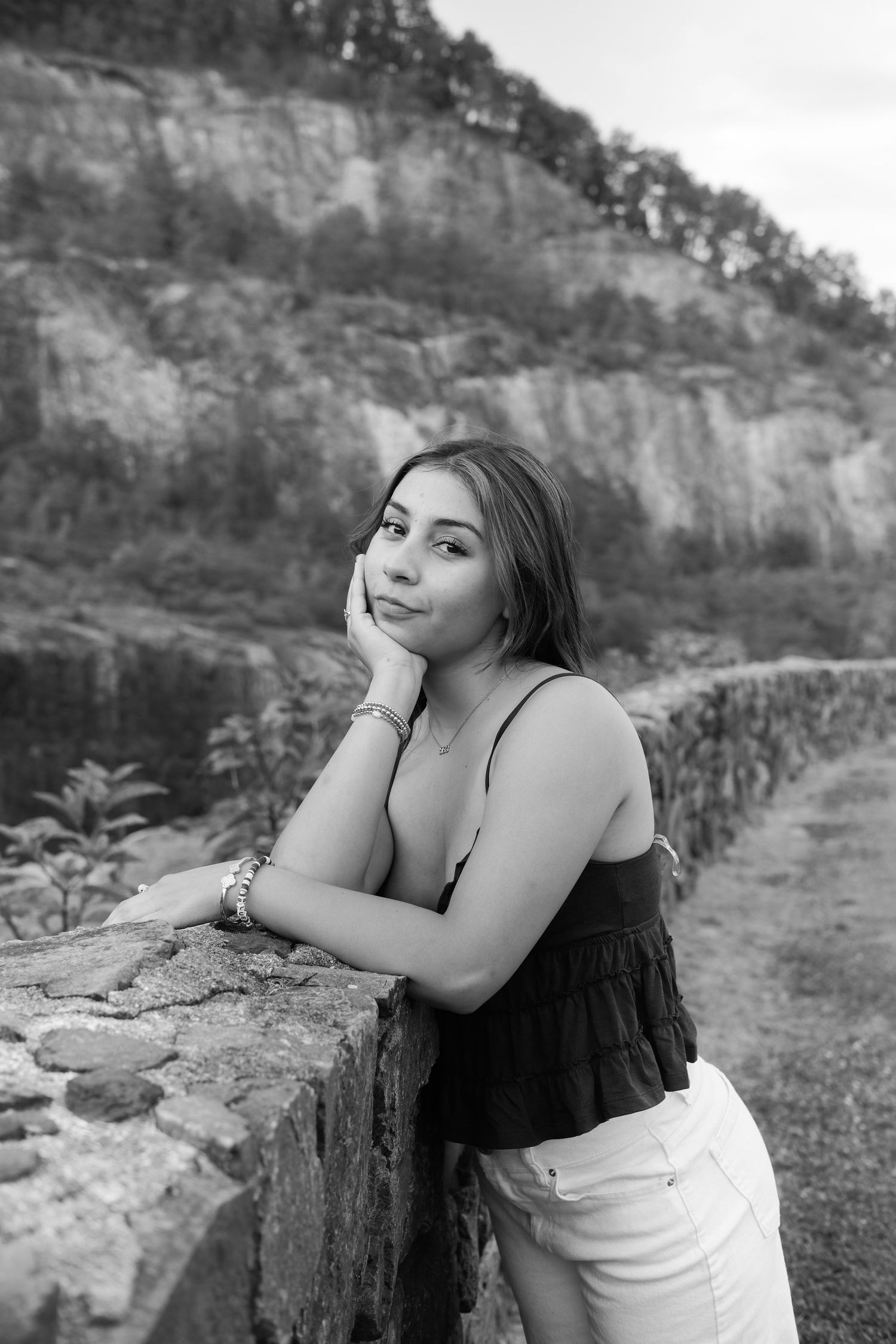 Black and white portrait of a young woman leaning on a stone wall with a rocky hillside in the background.