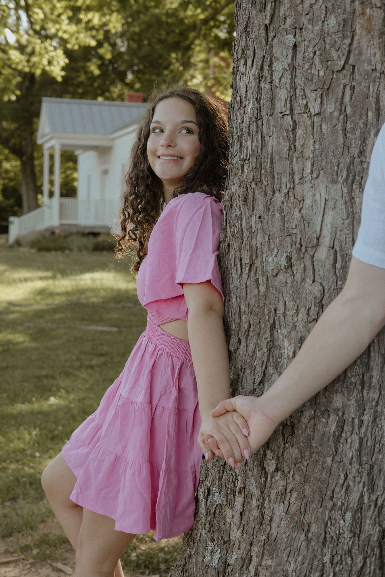 A smiling girl in a pink dress leans against a tree trunk, holding hands with someone in a sunny park.