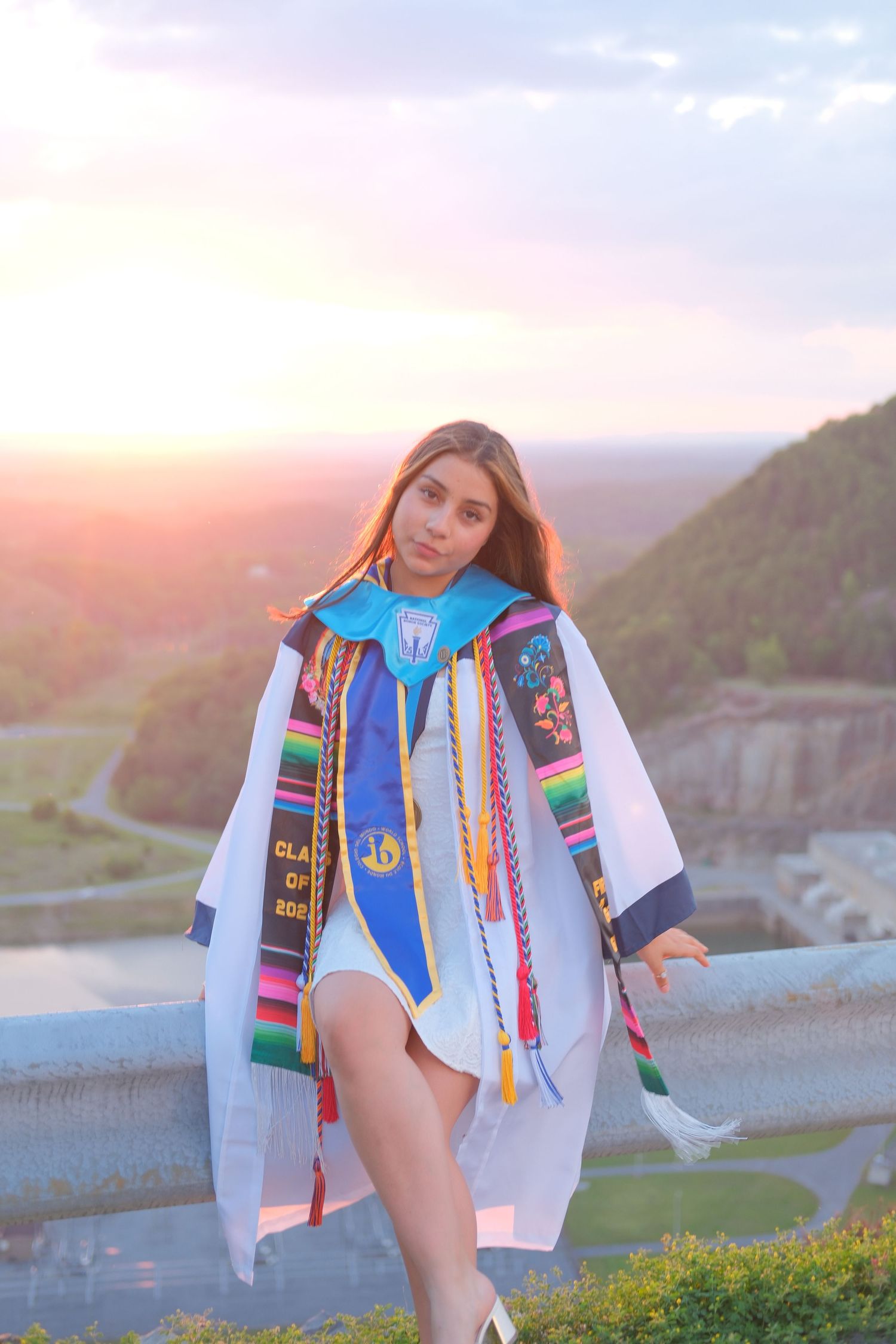 Young woman in graduation gown with colorful sashes poses at sunset with scenic mountain valley backdrop.