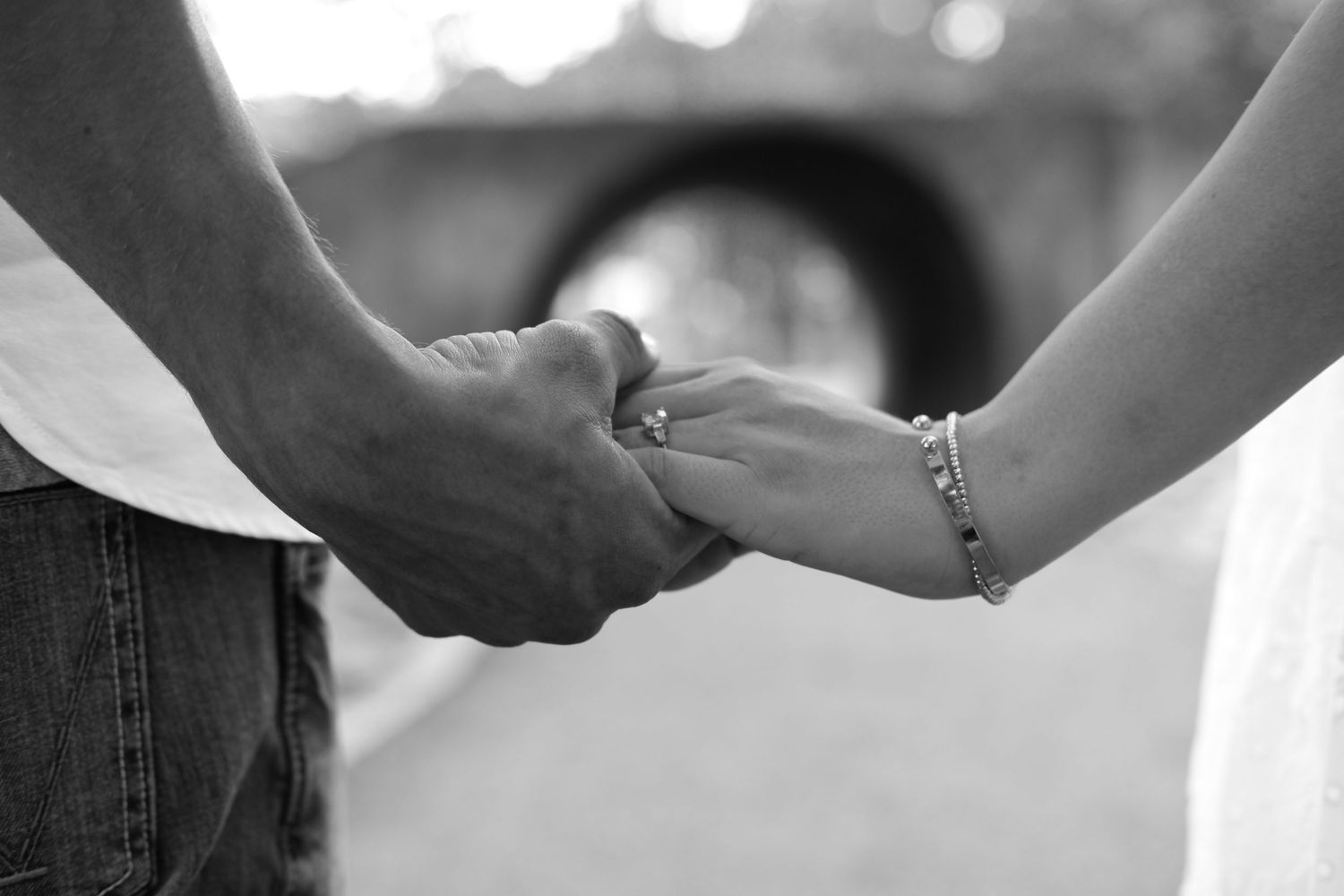 Black and white photo of a couple holding hands, woman wearing a delicate bracelet, romantic outdoor setting.