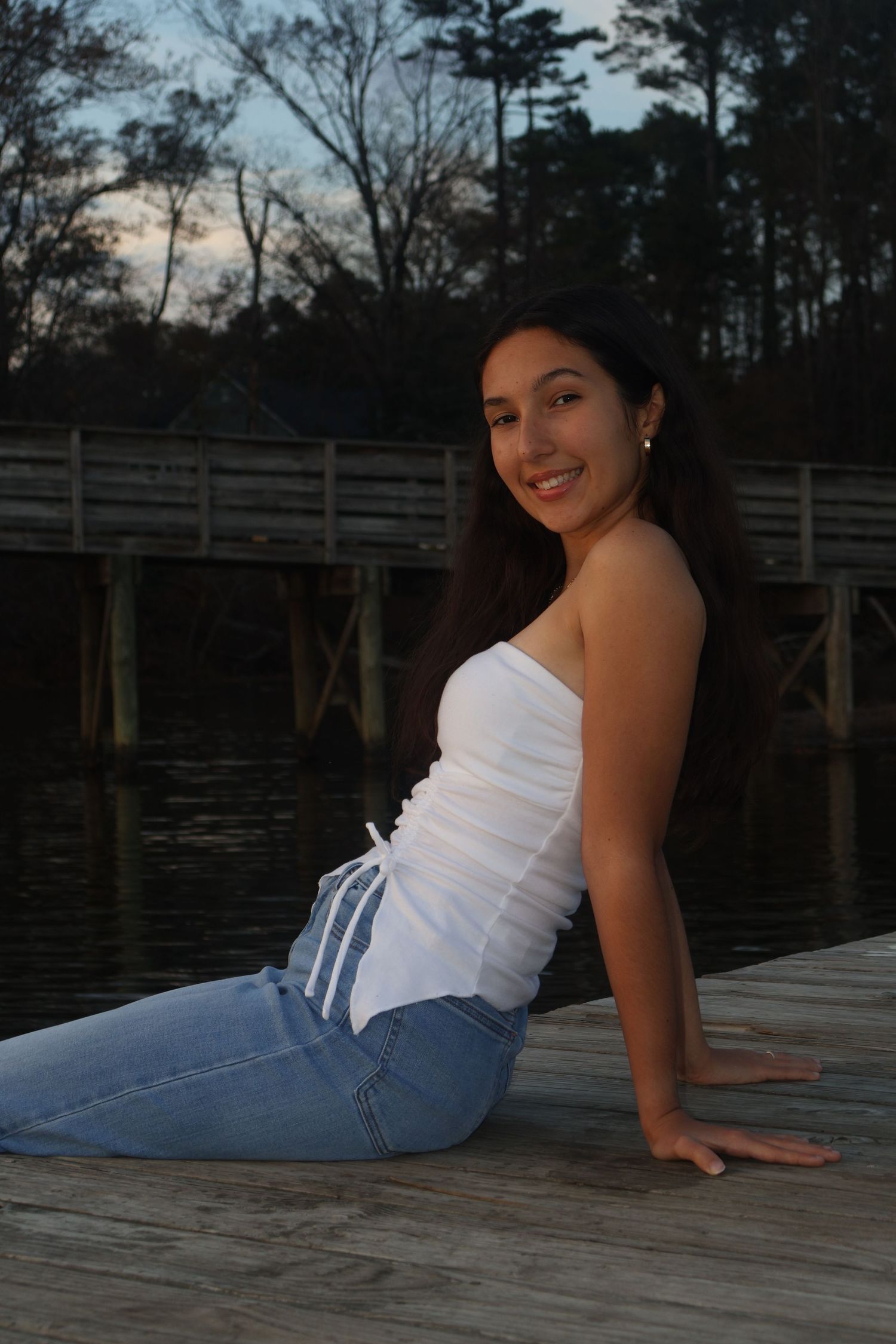 Young woman in white tube top and jeans sitting on wooden dock by water at dusk, smiling over shoulder.