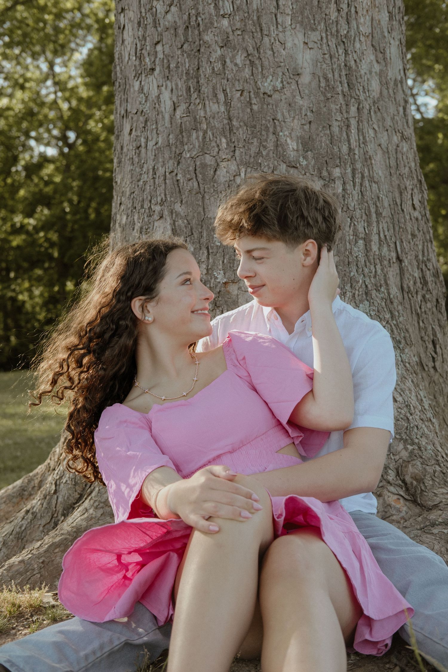 A young couple sits together against a large tree, smiling at each other, woman in pink dress, man in white shirt.