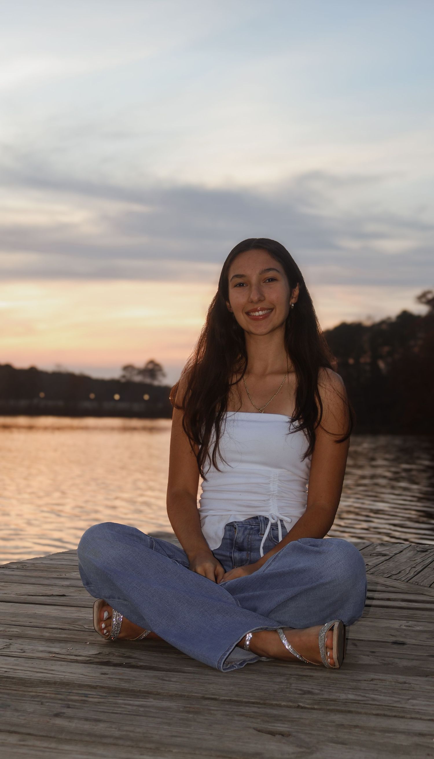 Young woman sitting cross-legged on a wooden dock at sunset, wearing white top and jeans.