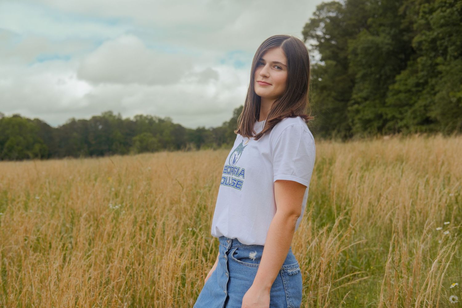 Smiling brunette teen in Georgia College t-shirt and jeans posing in golden field under cloudy sky.