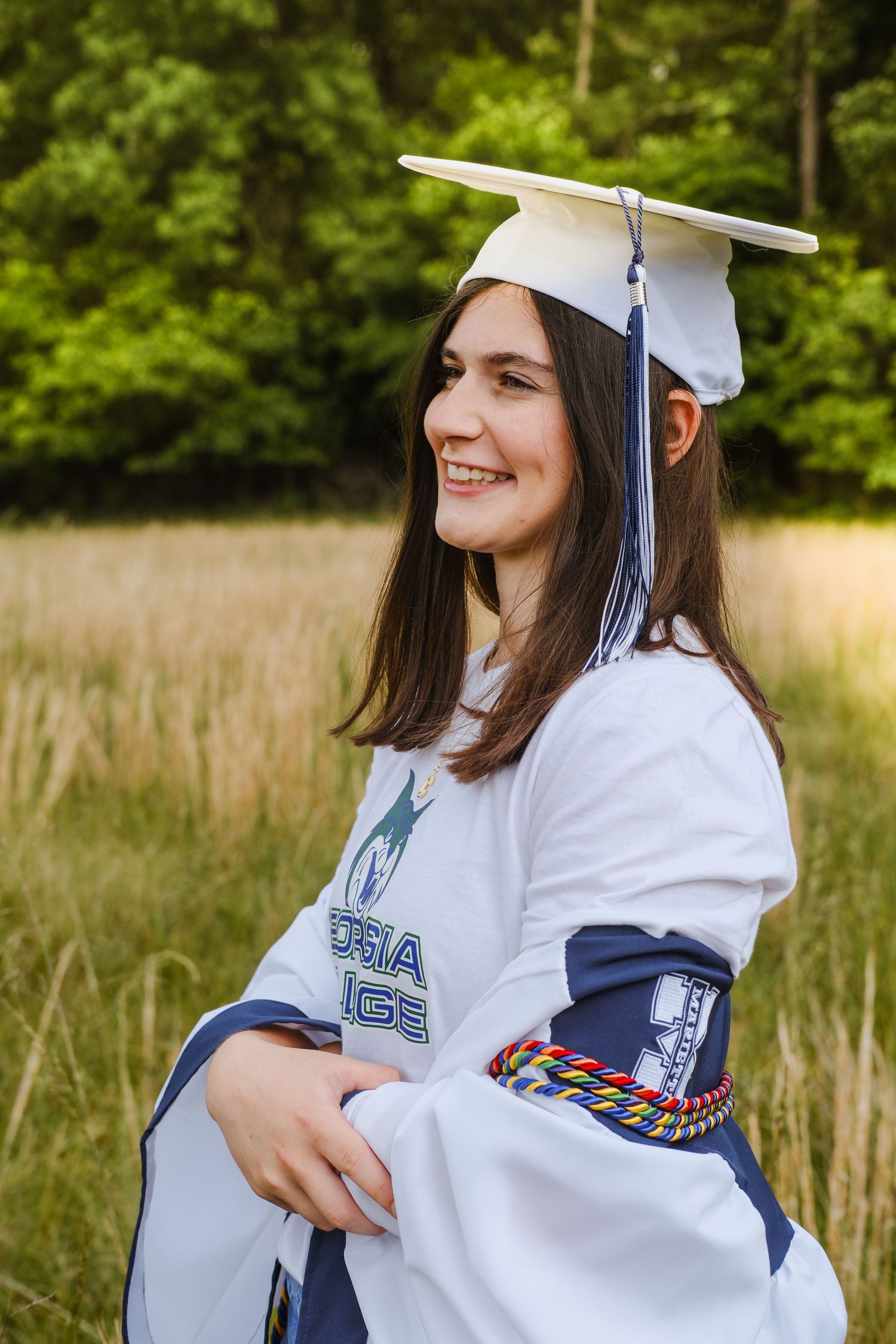 Smiling young woman in white graduation cap and gown with honor cords in a field.