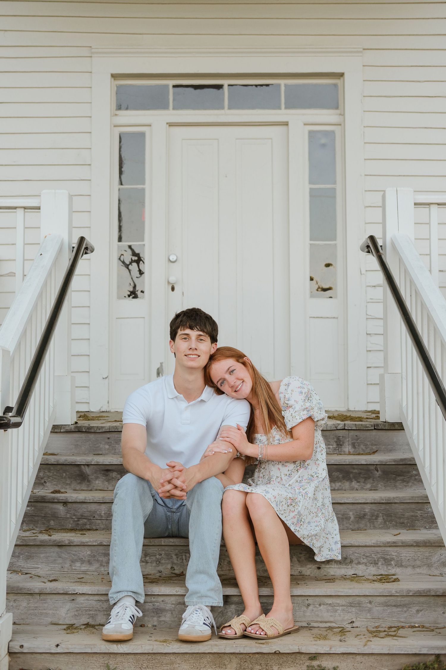 Young couple sitting on wooden steps of a white building, woman resting head on man's shoulder.