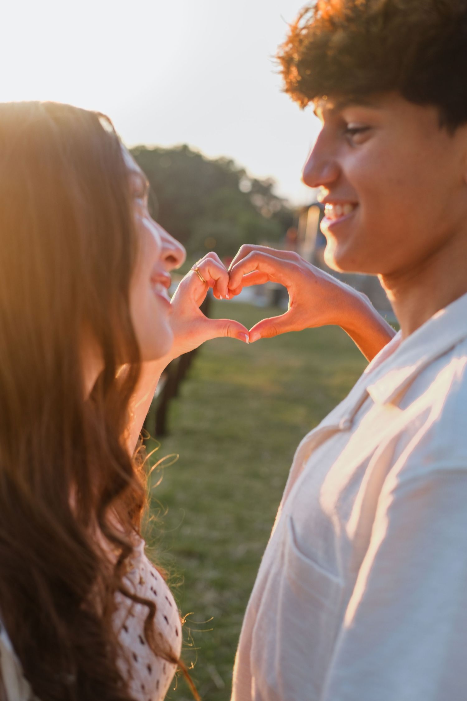 A young couple smiles while forming a heart shape with their hands at golden hour in a park.