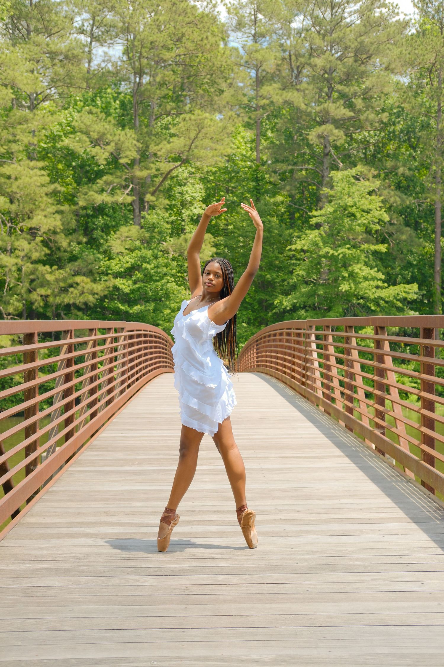 A ballerina in a white dress performs on pointe on a wooden bridge surrounded by lush green trees.