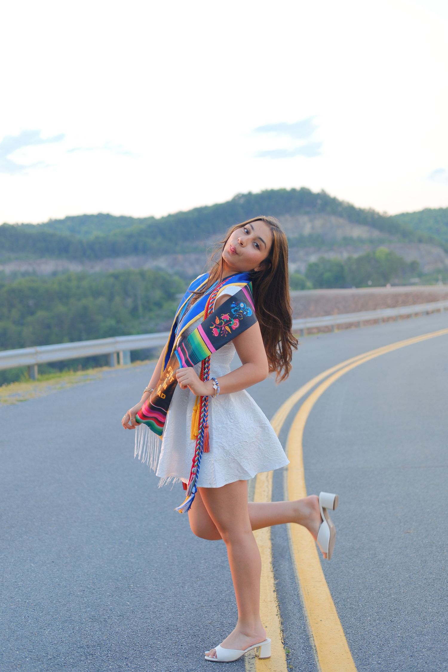 Young woman in white dress and colorful serape shawl poses on yellow center line of scenic mountain road.